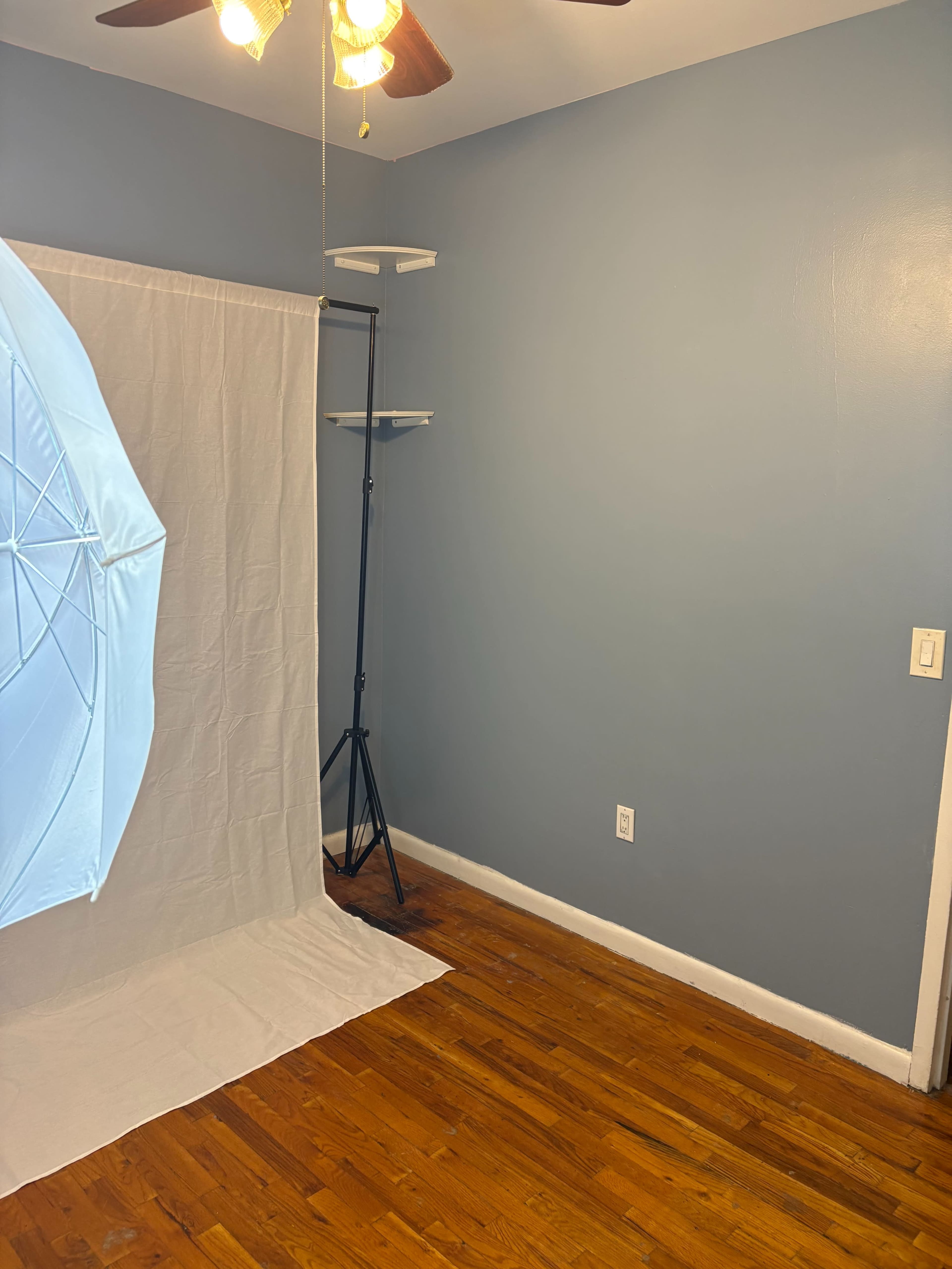 The image shows a small room with blue walls, a wooden floor, a lighting umbrella set up on the left, and a plain white backdrop.