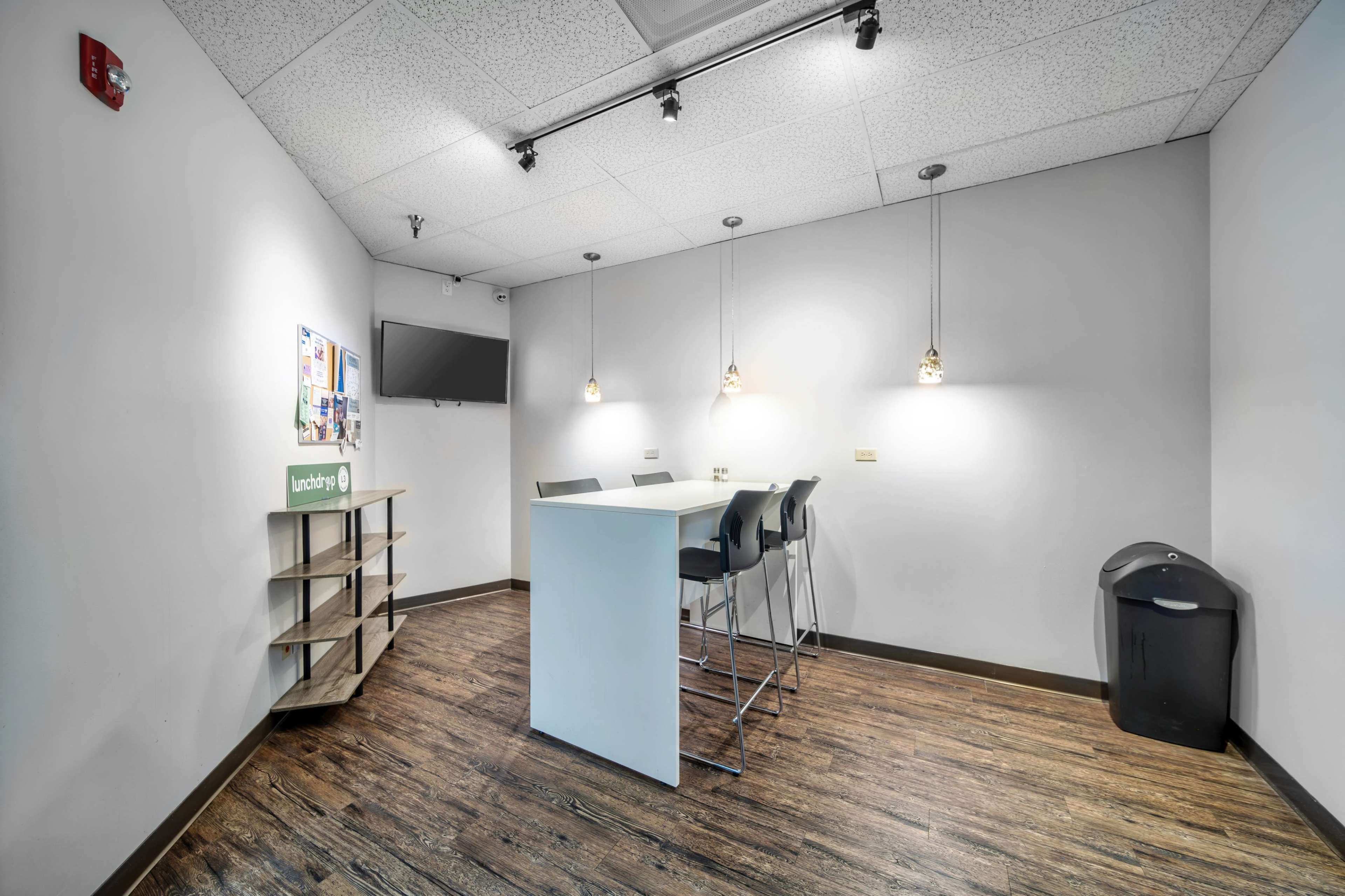 The image shows a modern room with a small table and high stools, a television mounted on the wall, and a storage shelf on one side.