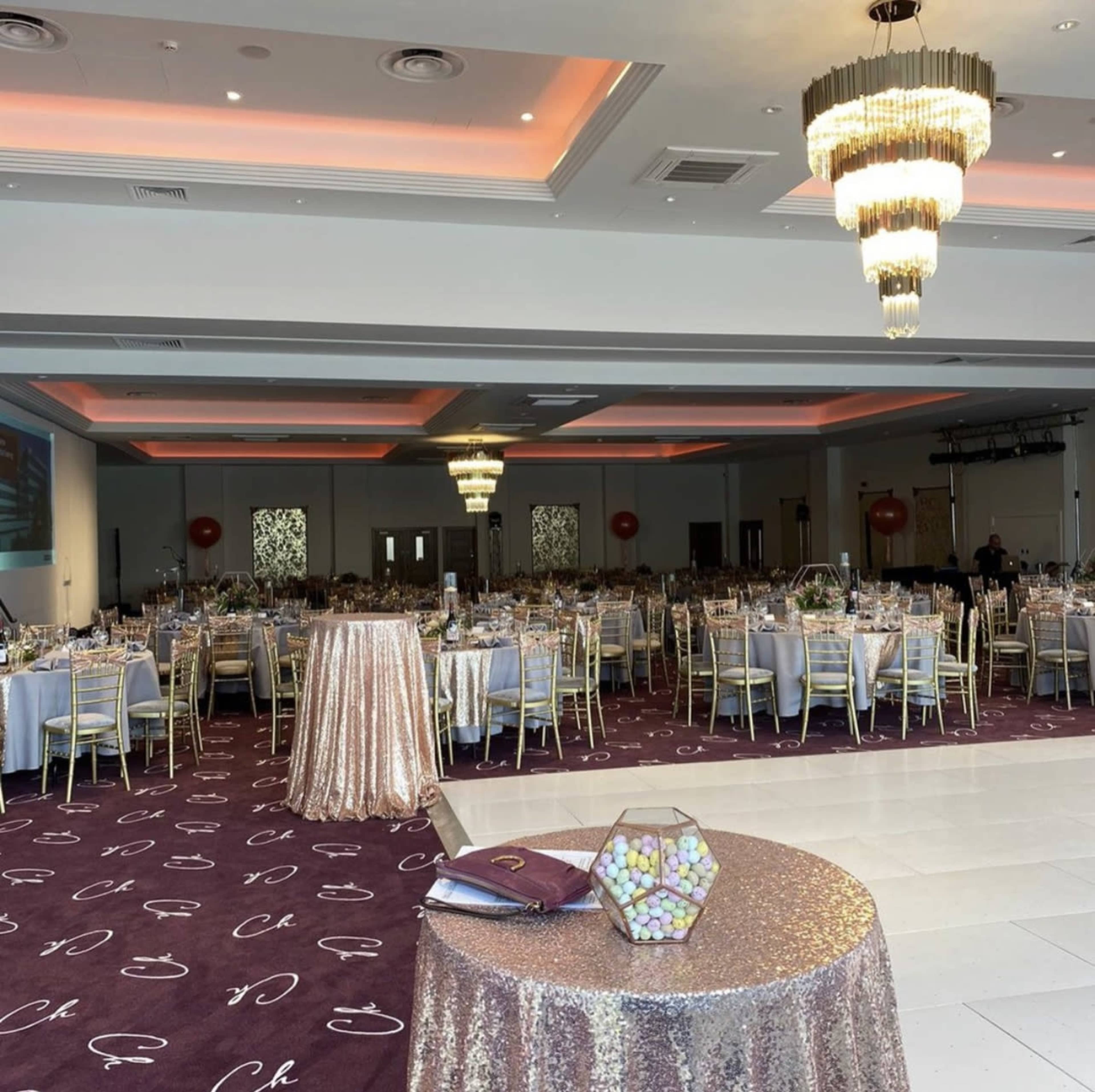The image shows a spacious banquet hall set up for an event, featuring tables with golden chairs, elegant table settings, and large chandeliers overhead.