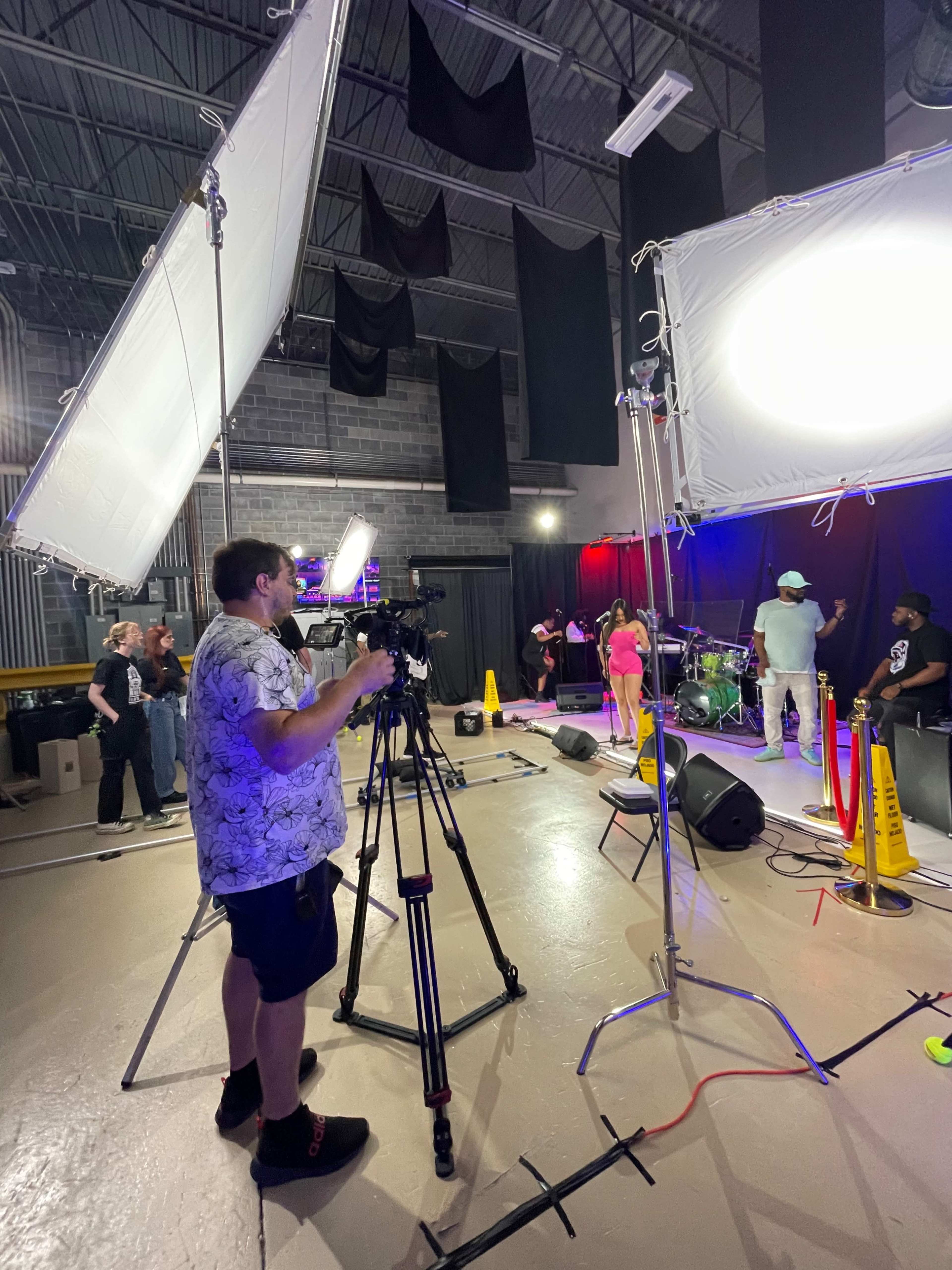 A person operates a camera on a tripod in a dimly lit studio where musicians are performing on stage.
