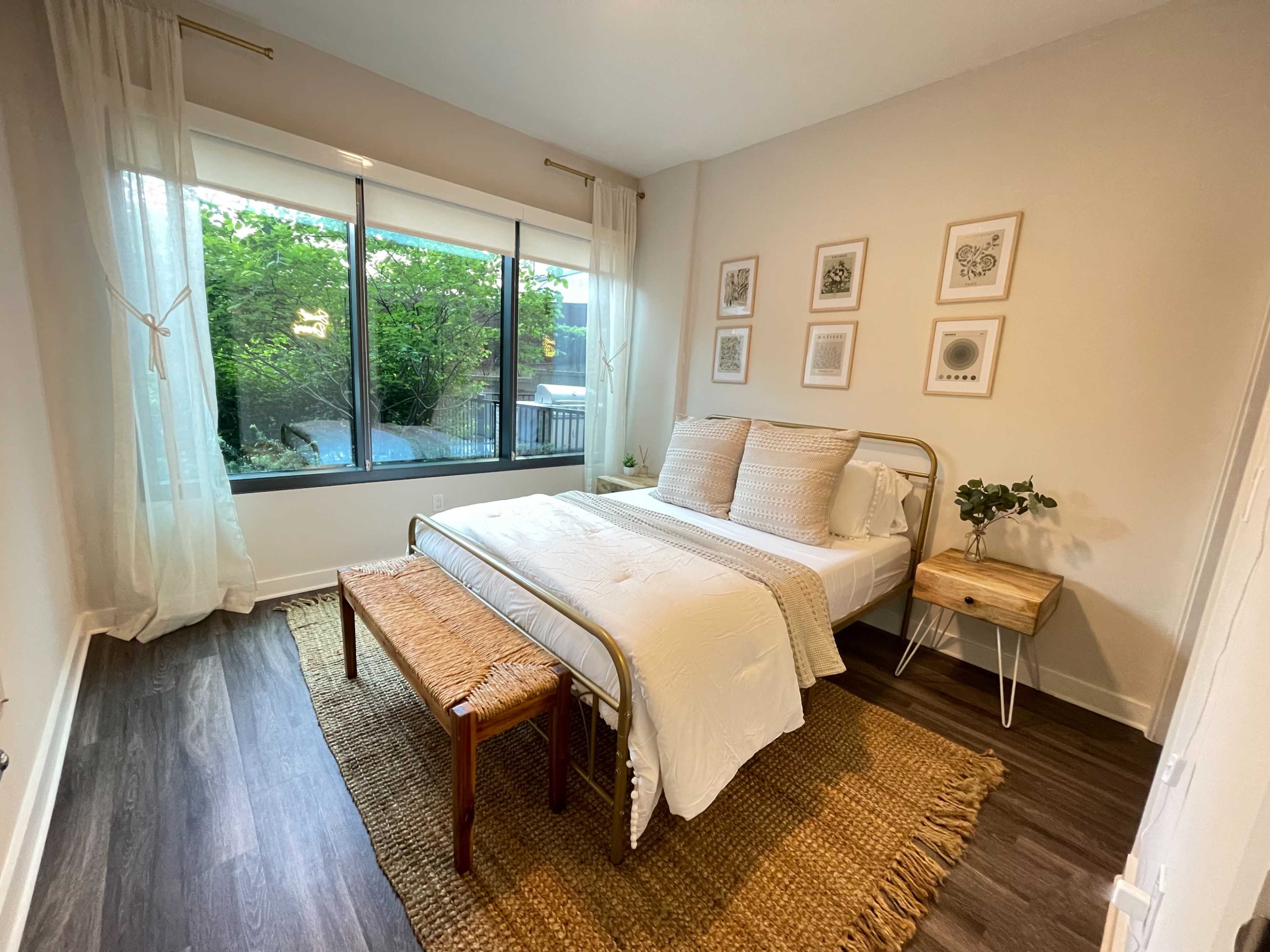 A neatly arranged bedroom features a bed with white bedding, a bench at the foot, and large windows allowing natural light and views of greenery.