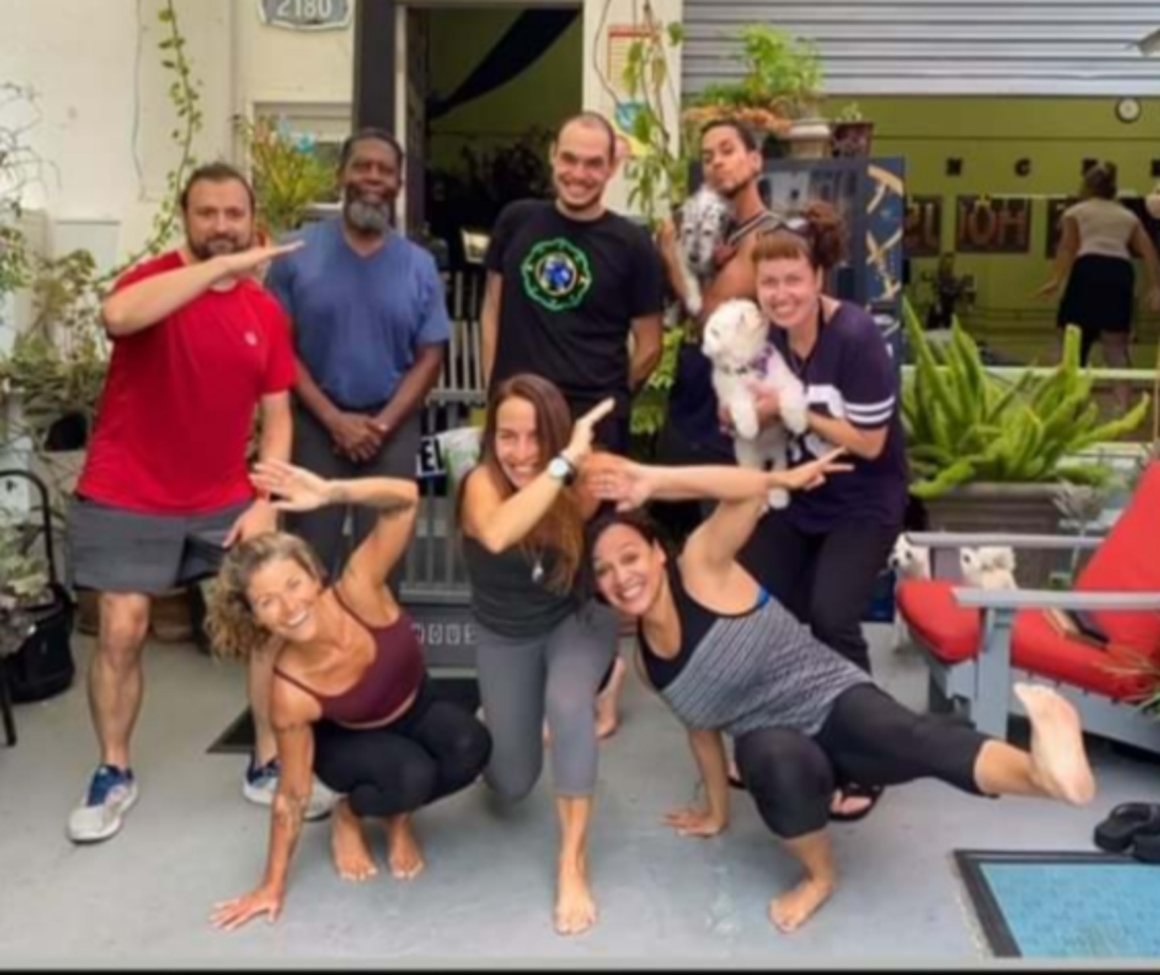 A group of eight people poses together in a yoga studio, with some performing various yoga poses and holding a small dog.