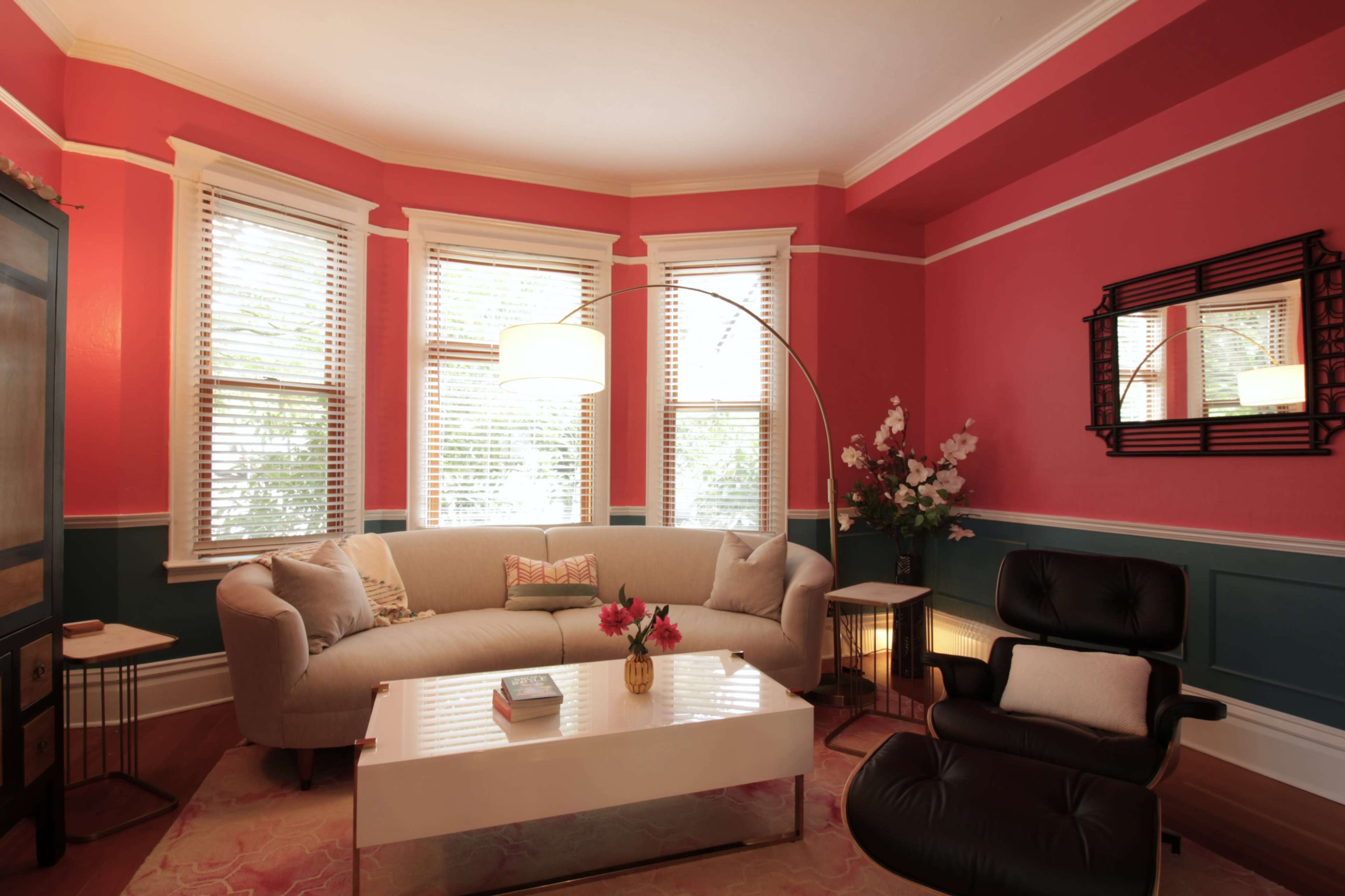 The living room features a curved sofa, a round floor lamp, and large windows with wooden blinds, all set against a backdrop of pink and teal walls.