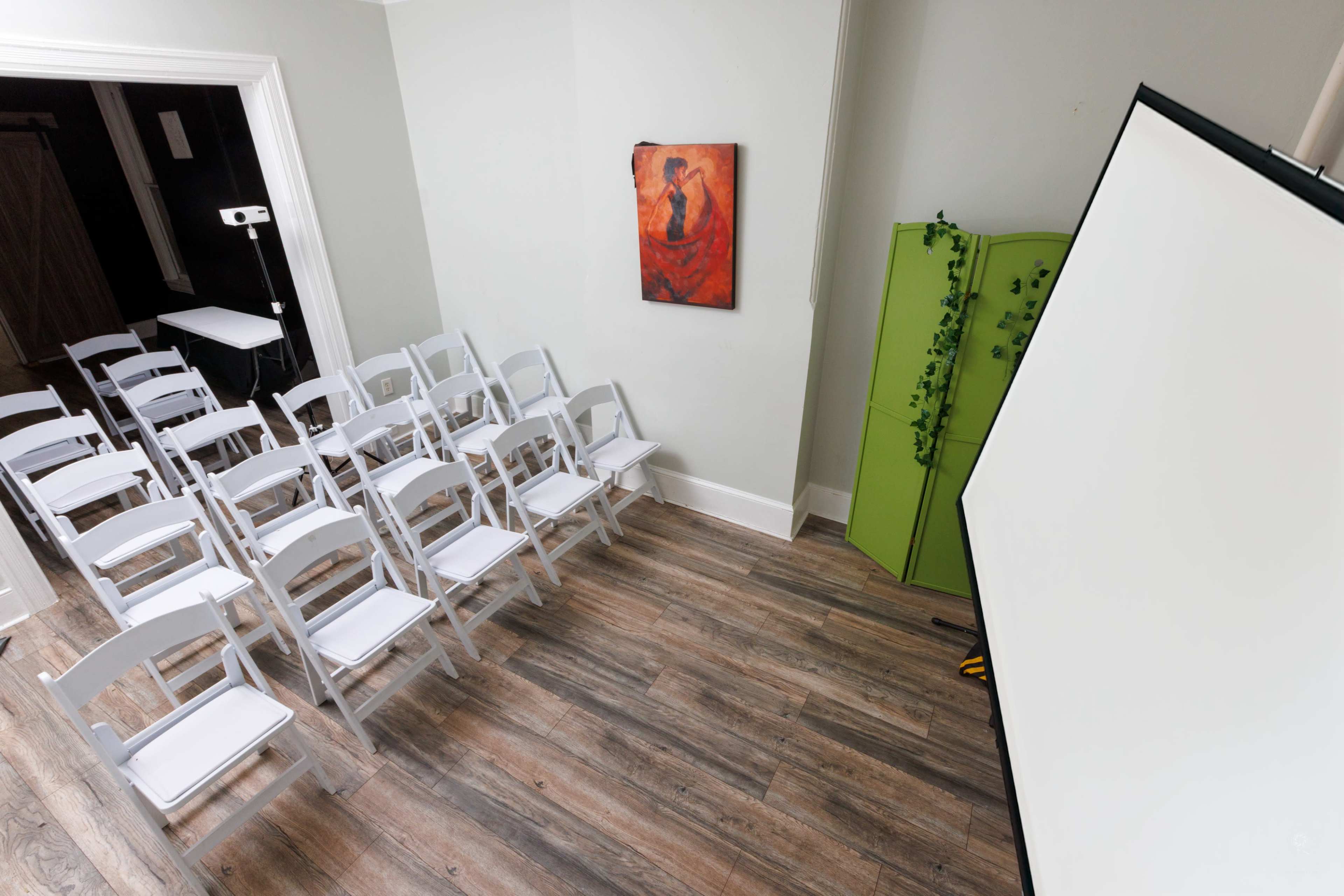 The image shows a small room arranged with white chairs facing a blank projection screen, accompanied by a painted artwork on the wall and a green folding partition.
