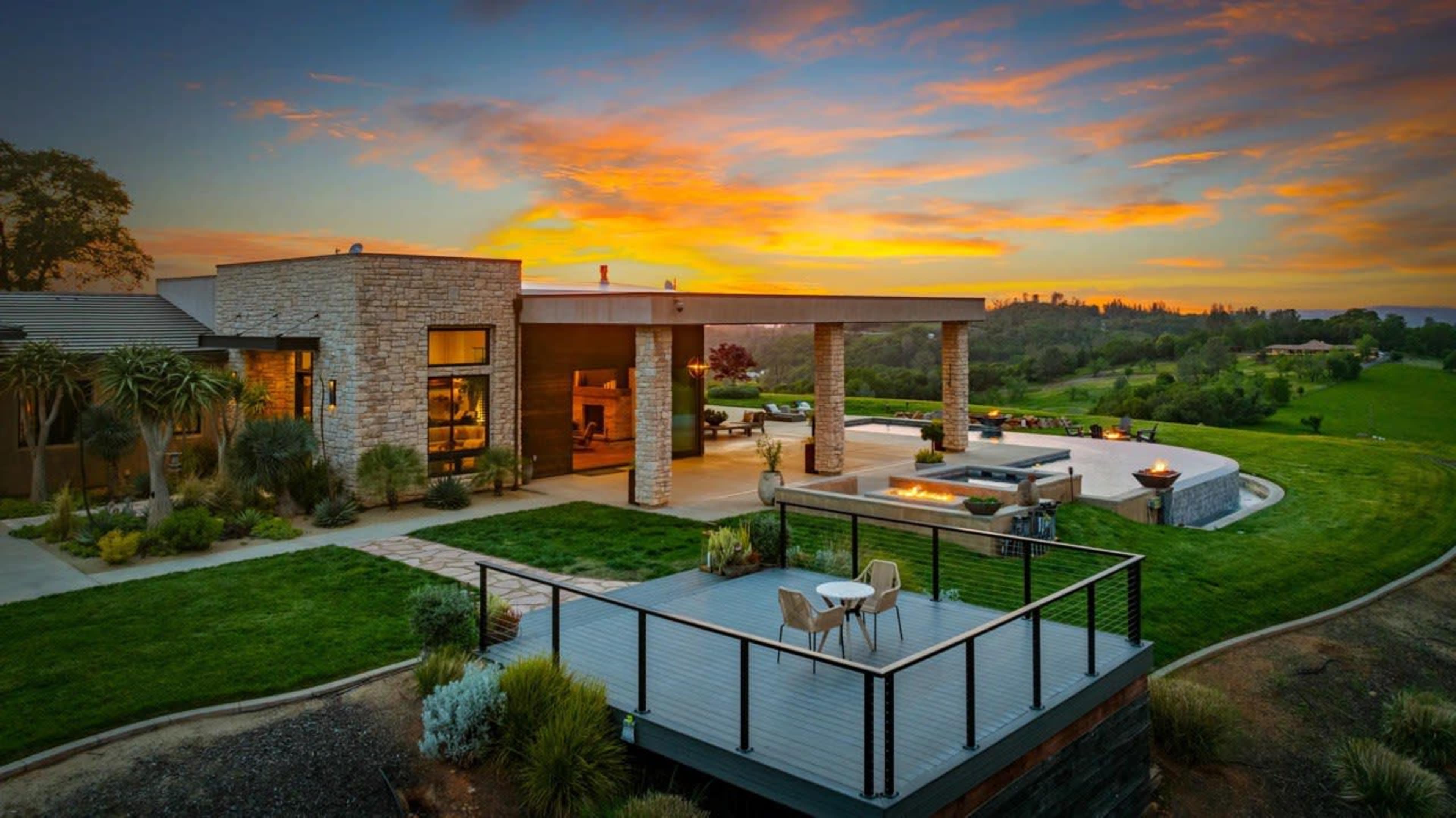 The image shows a modern house with large windows and a patio overlooking a lush green landscape during sunset.