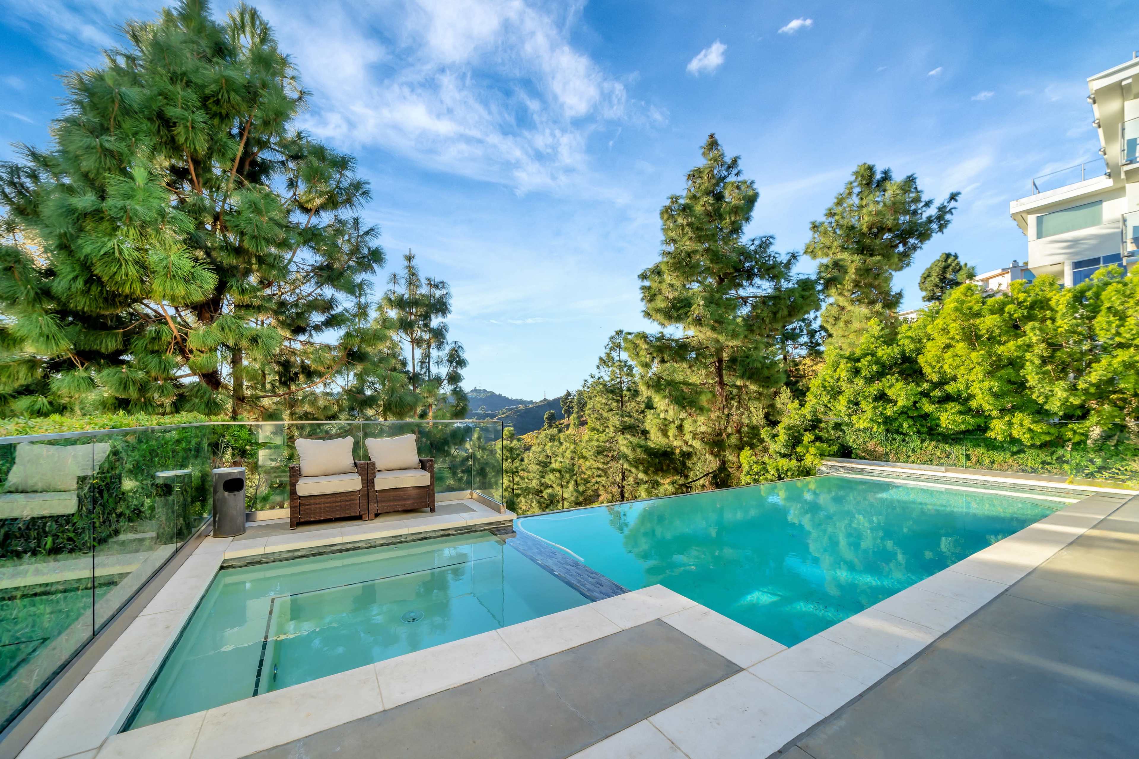 The image shows a modern infinity pool overlooking a mountainous landscape, surrounded by tall trees and clear blue skies.