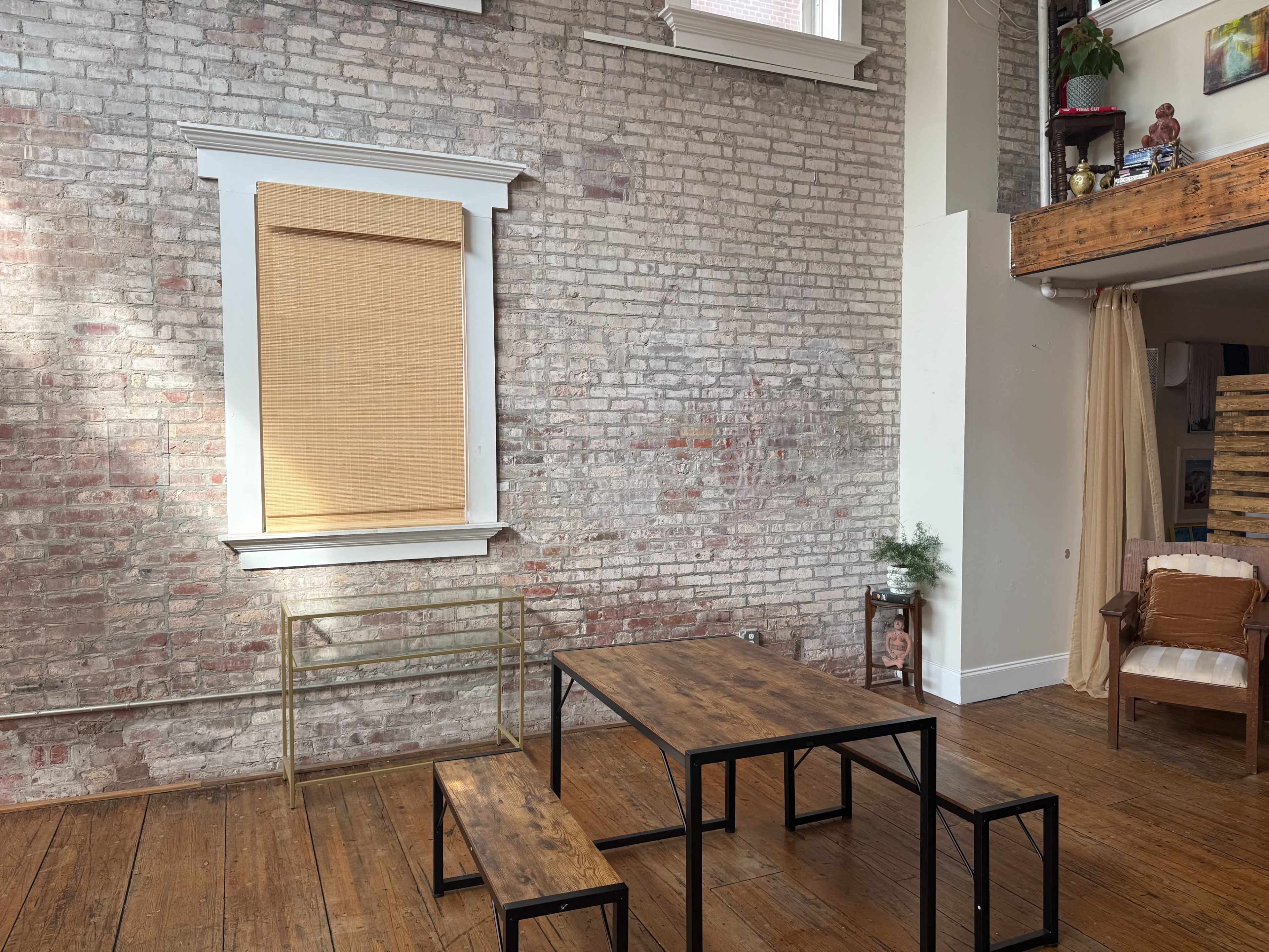 The image shows a spacious room with exposed brick walls, a wooden floor, a large window covered by a bamboo shade, and a simple wooden table with benches.
