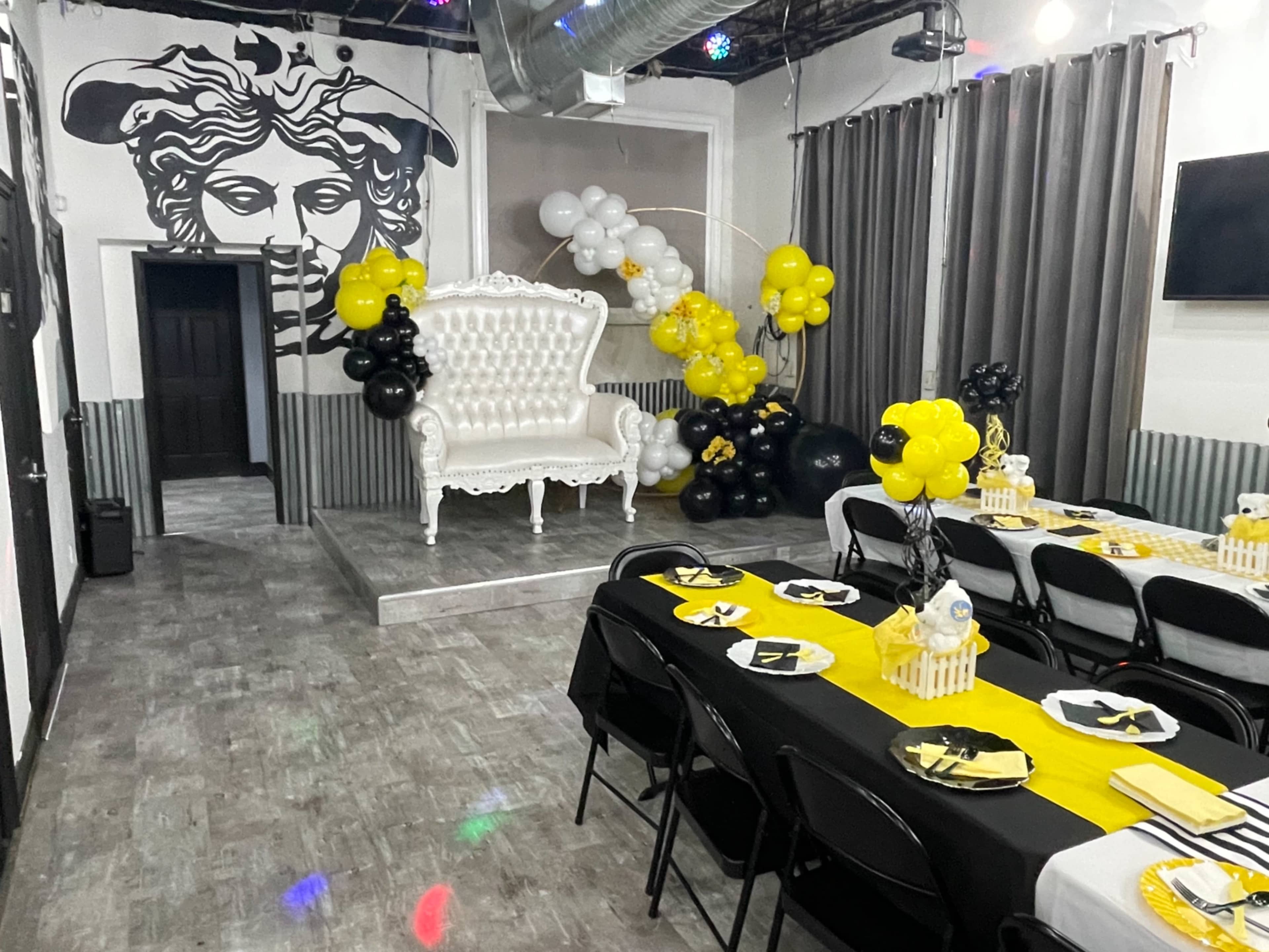 A decorated event space featuring a white ornate sofa, black and yellow balloons, and tables set with black tablecloths and yellow accents.
