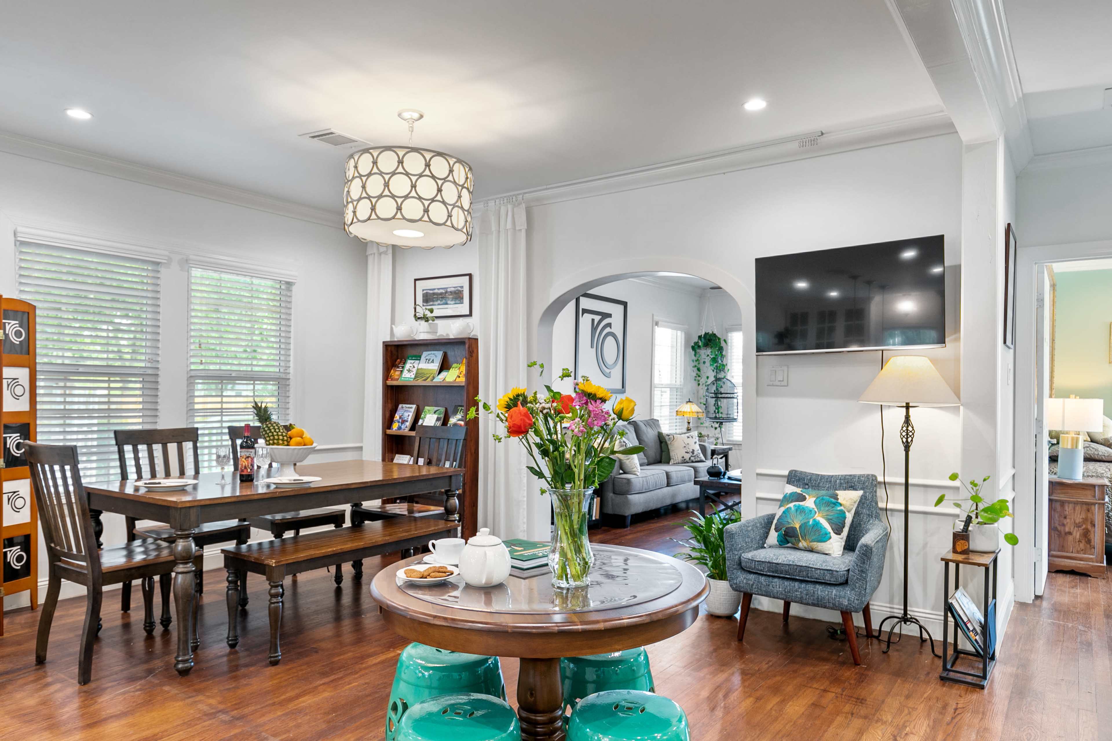 A bright living space features a dining area with a wooden table, a seating area with a television, and colorful flowers on the table.