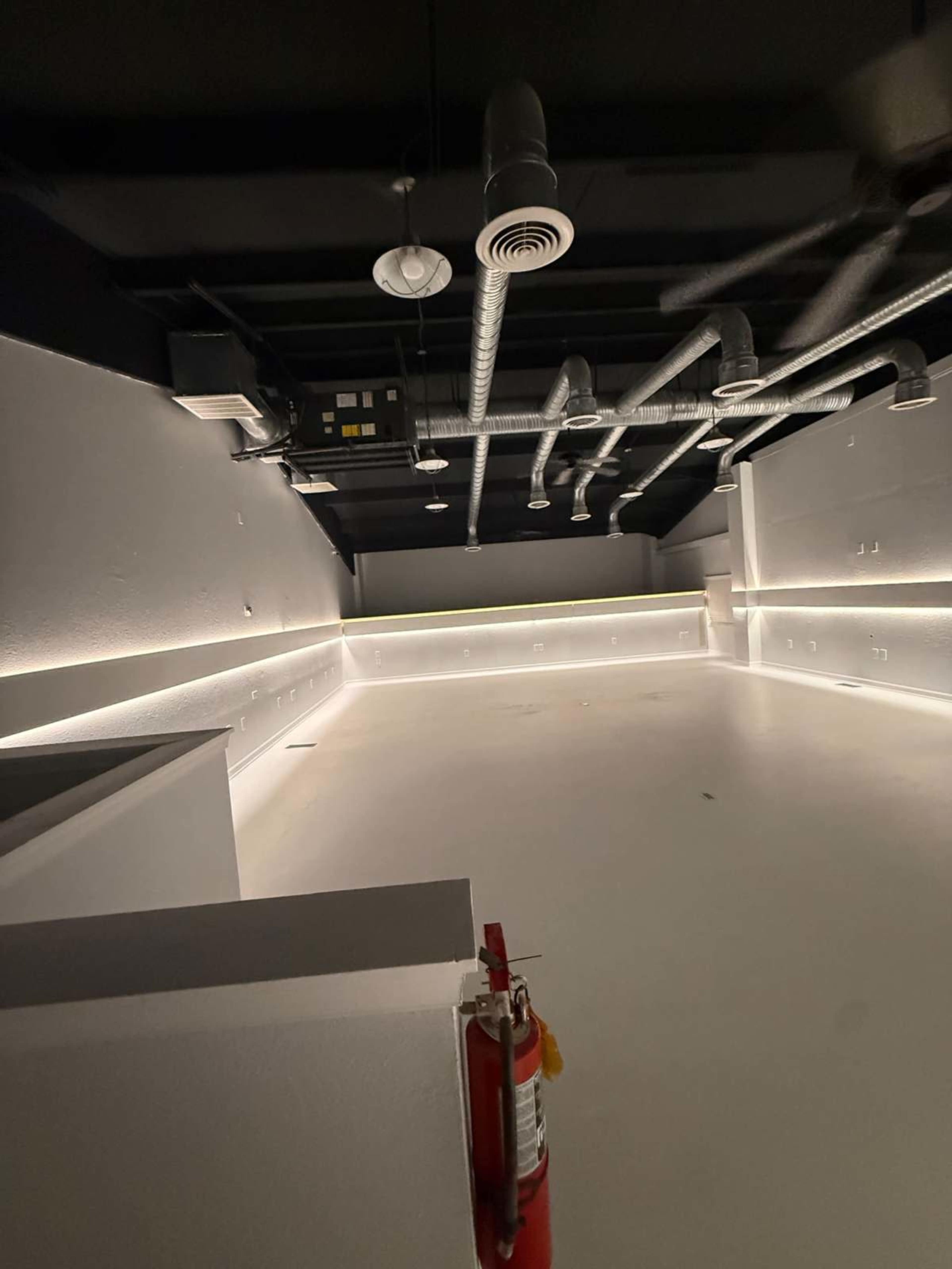 The image shows an empty room with white walls and a light-colored floor, featuring overhead ventilation ducts and a fire extinguisher in the foreground.