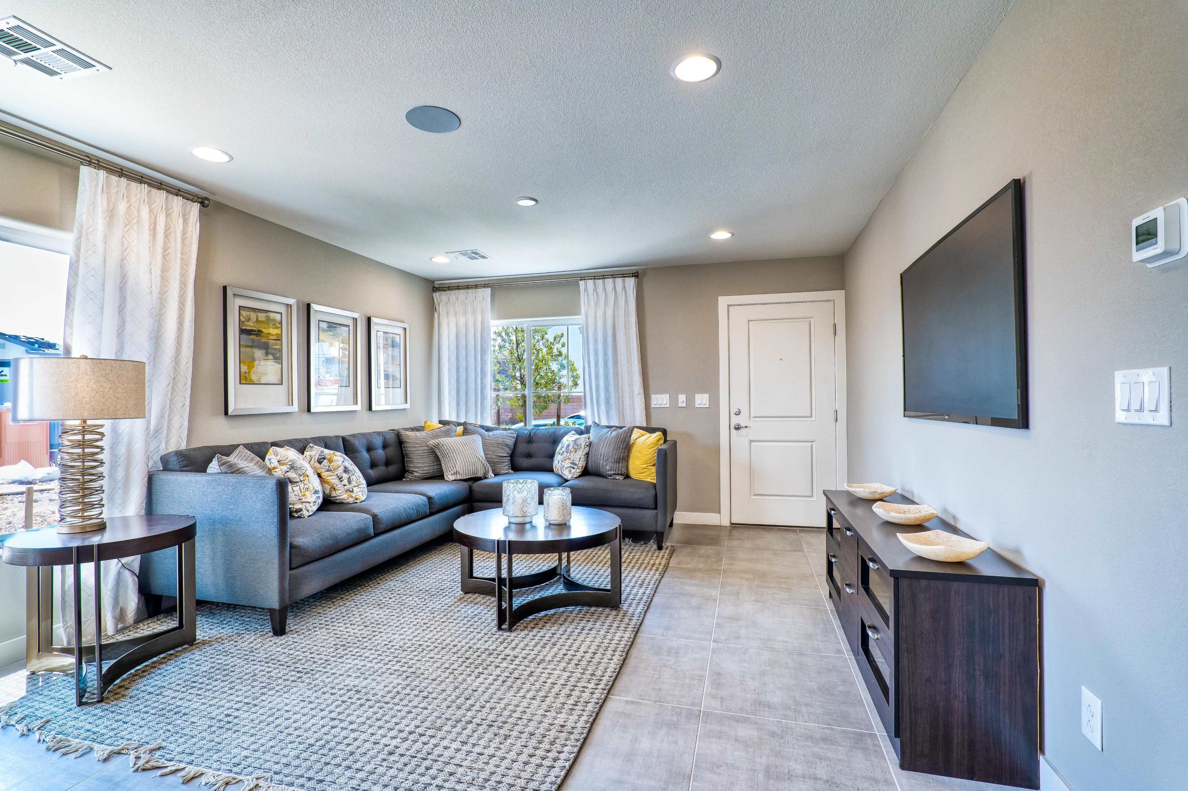 A modern living room features a sectional sofa, a coffee table, and a television mounted on the wall, with windows allowing natural light to enter.