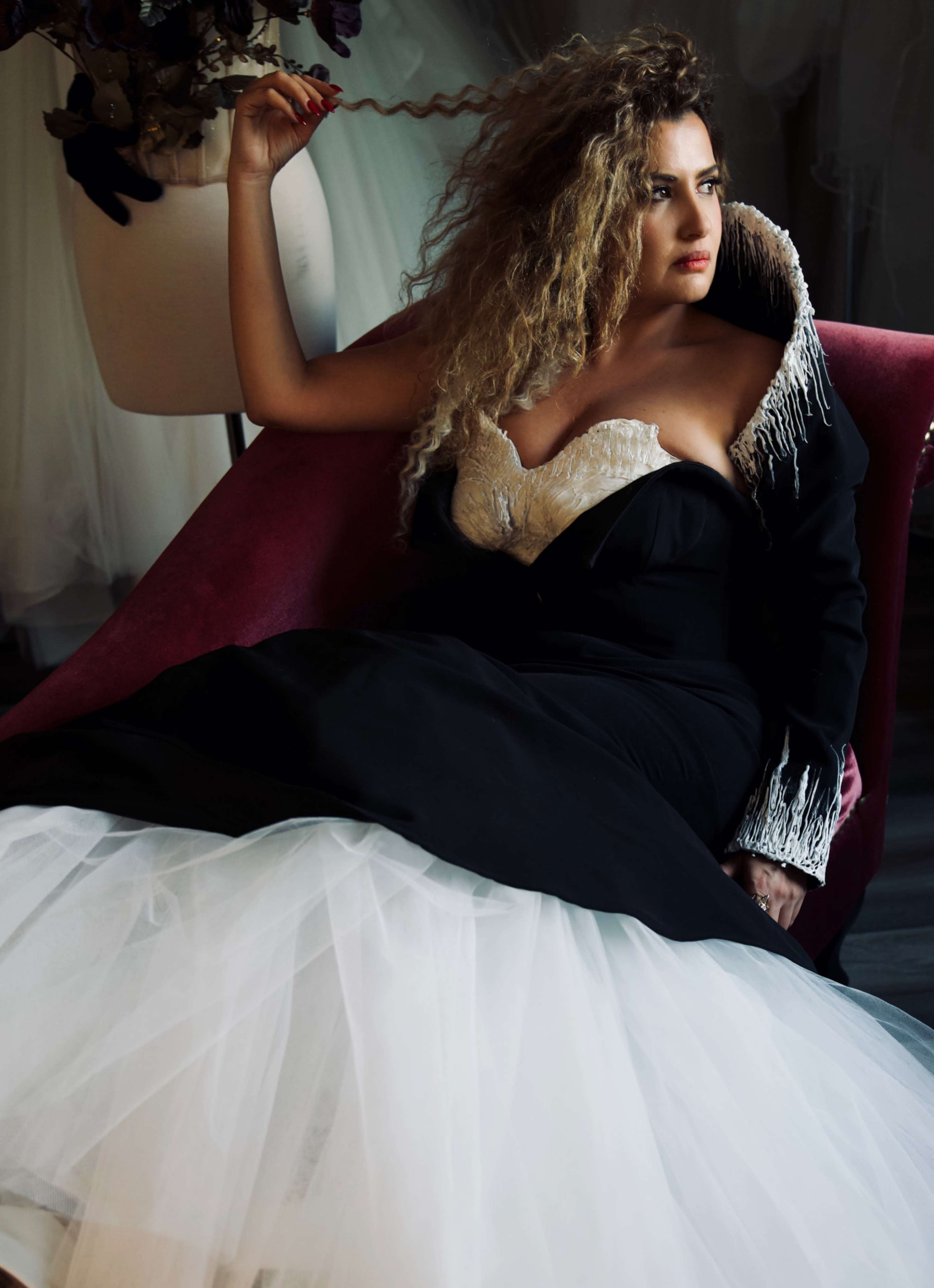 A woman sits on a pink chair wearing a black gown with a white corset and a long, flowing tulle skirt.