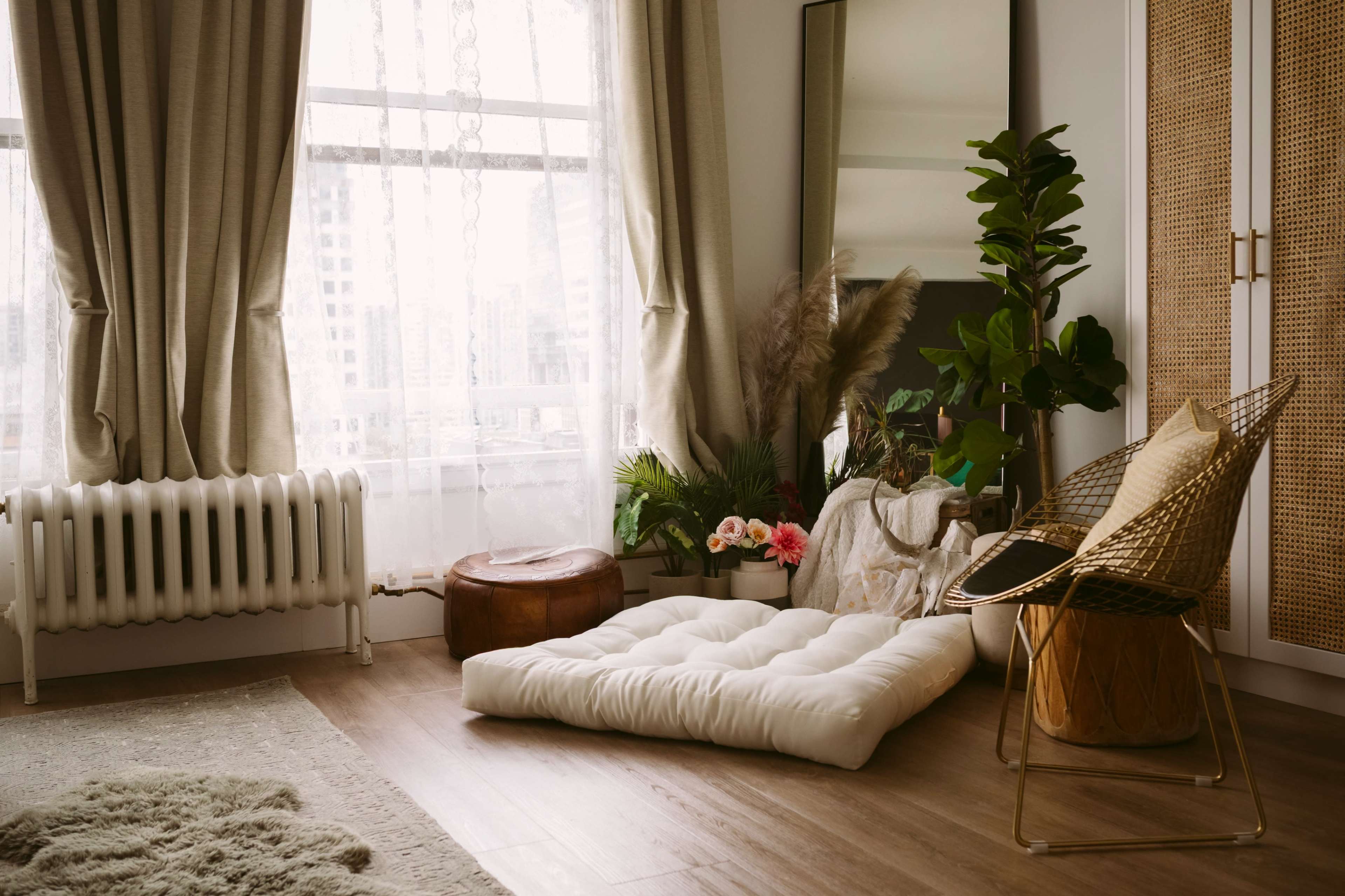 A minimalist living space features a large window with sheer curtains, a floor cushion, a decorative chair, a wooden stool, and various plants.