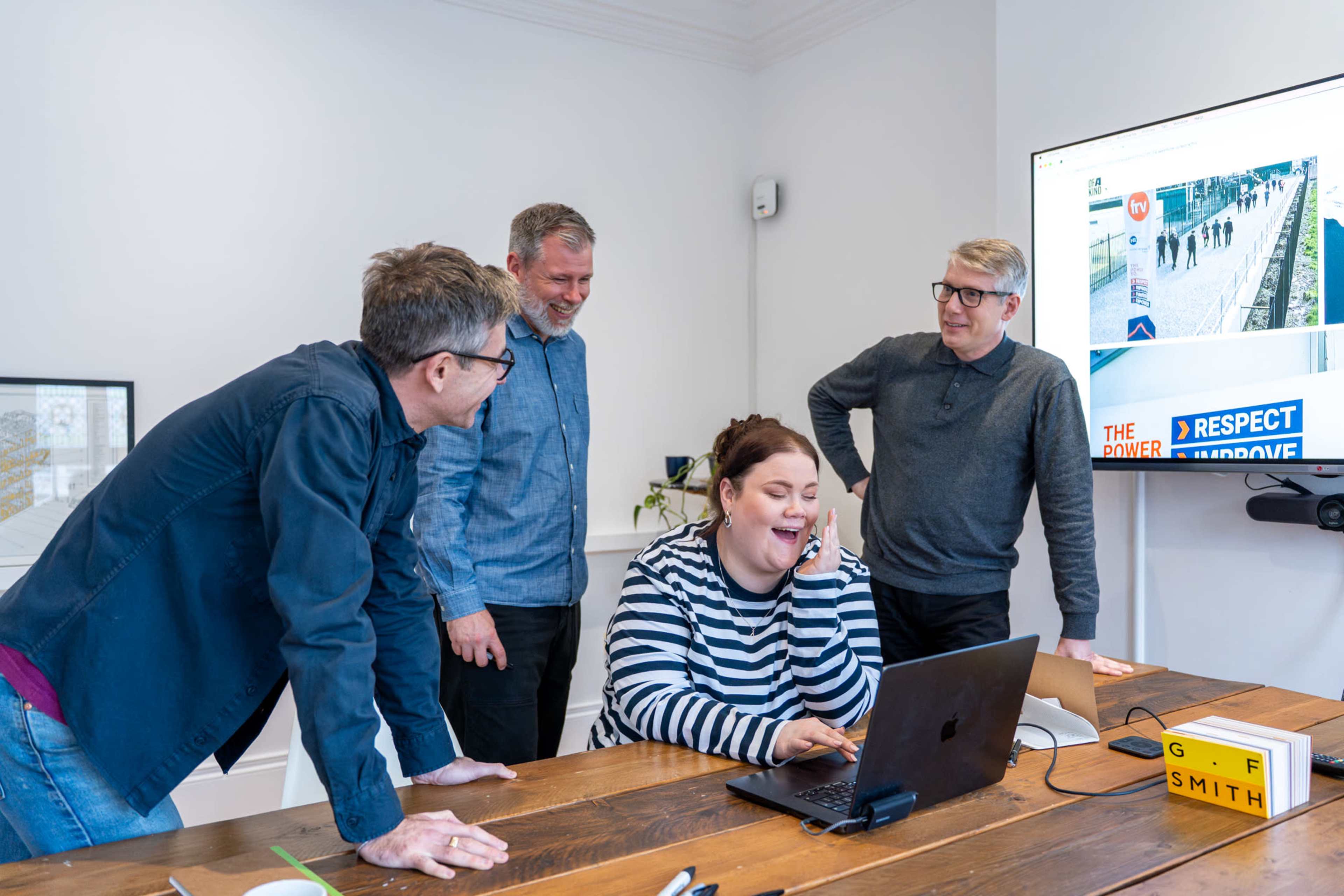 Four individuals are gathered around a wooden table, engaging with a laptop while a screen in the background displays a photo and text related to a presentation.