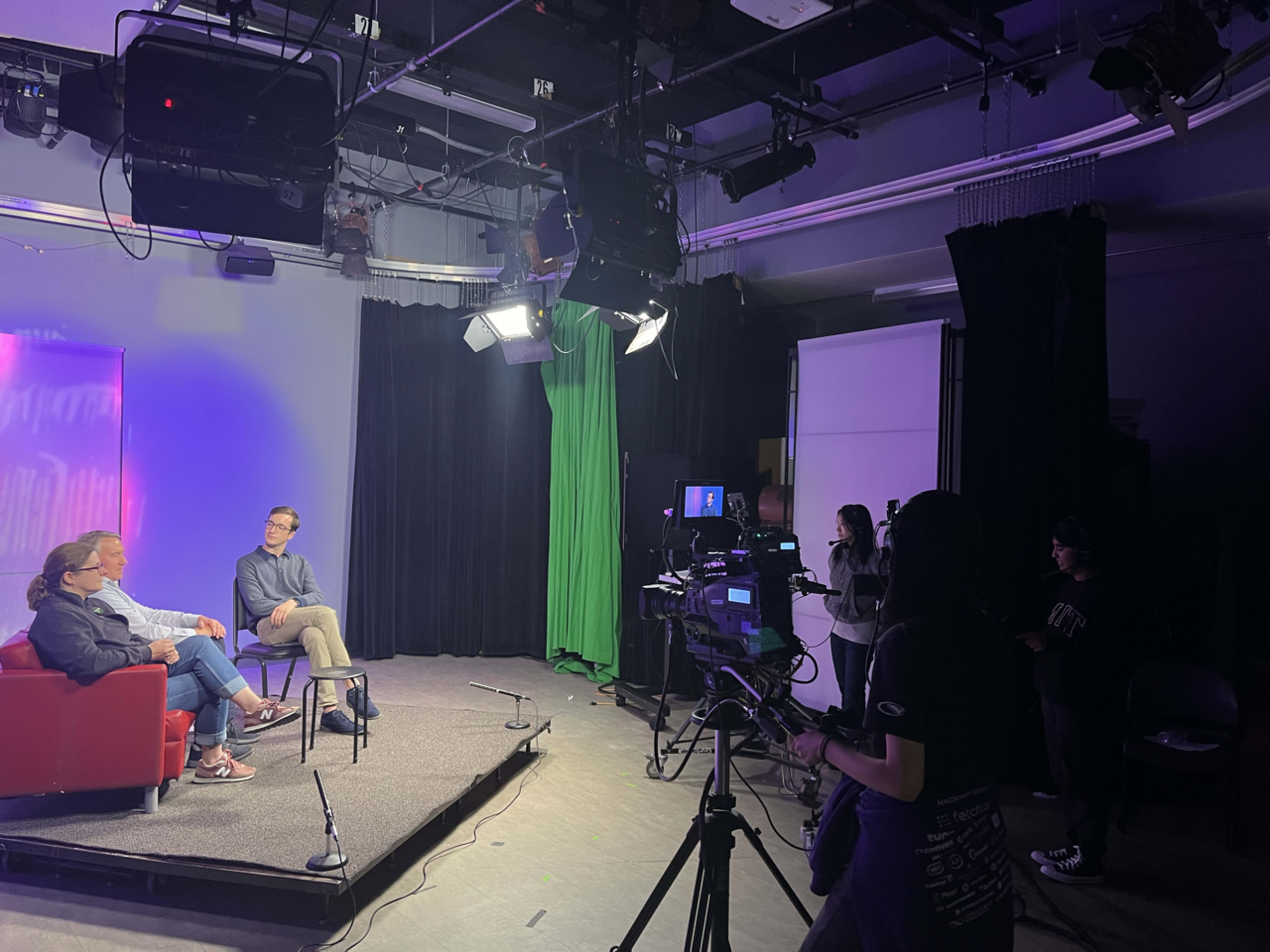 The image shows a television studio setup with two people seated on a red couch and several cameras and lights positioned around them.