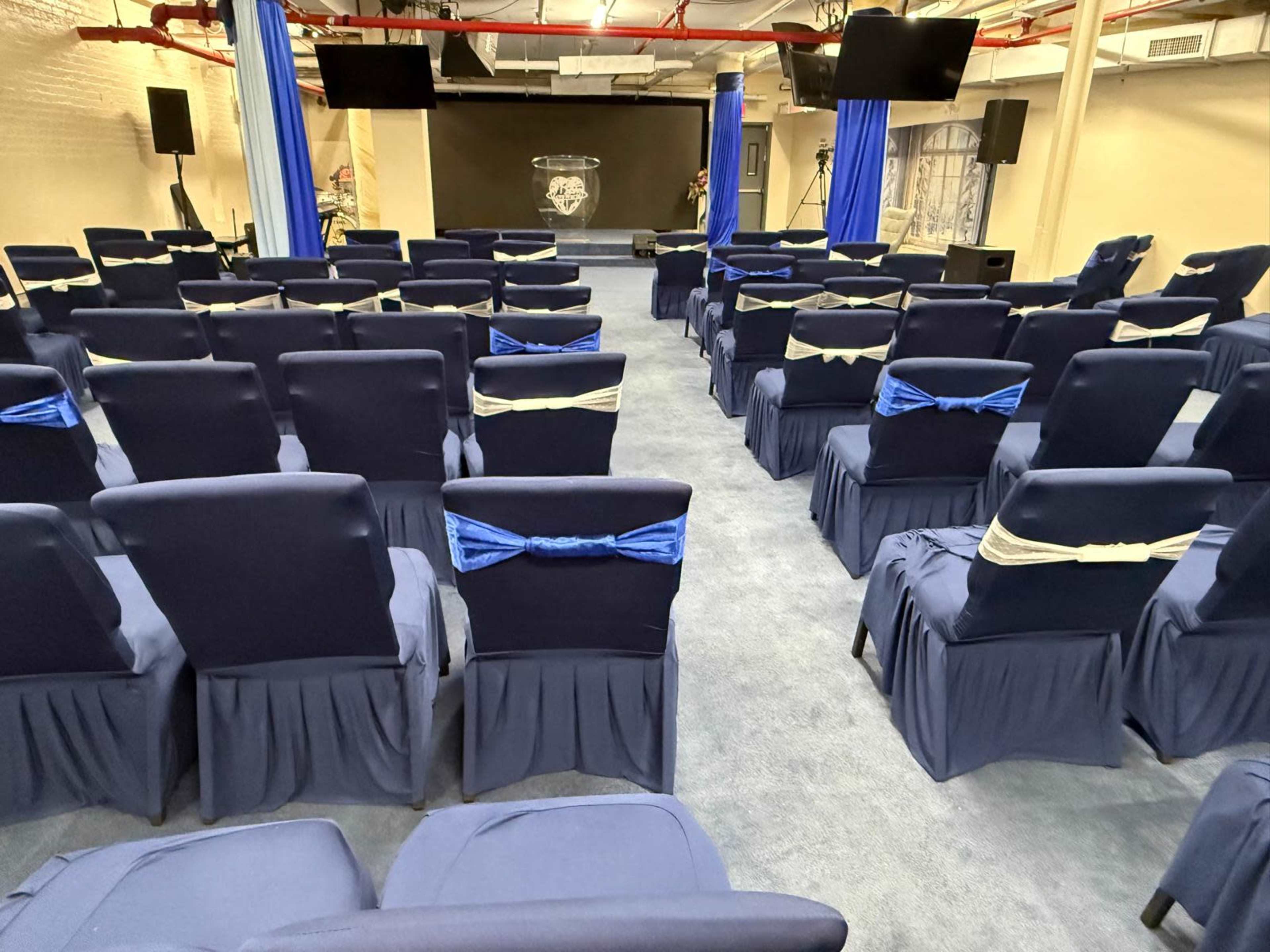 The image shows an indoor event space with rows of navy blue chairs adorned with light blue bows, arranged facing a stage with a black backdrop and audio-visual equipment.