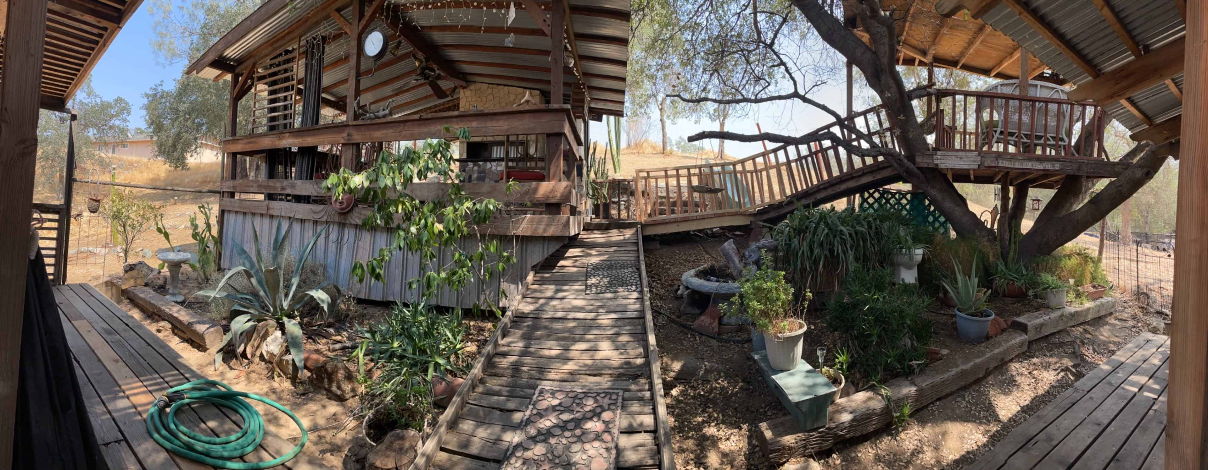A wooden deck and garden area with various plants, leading to a house elevated on stilts under a sunny sky.