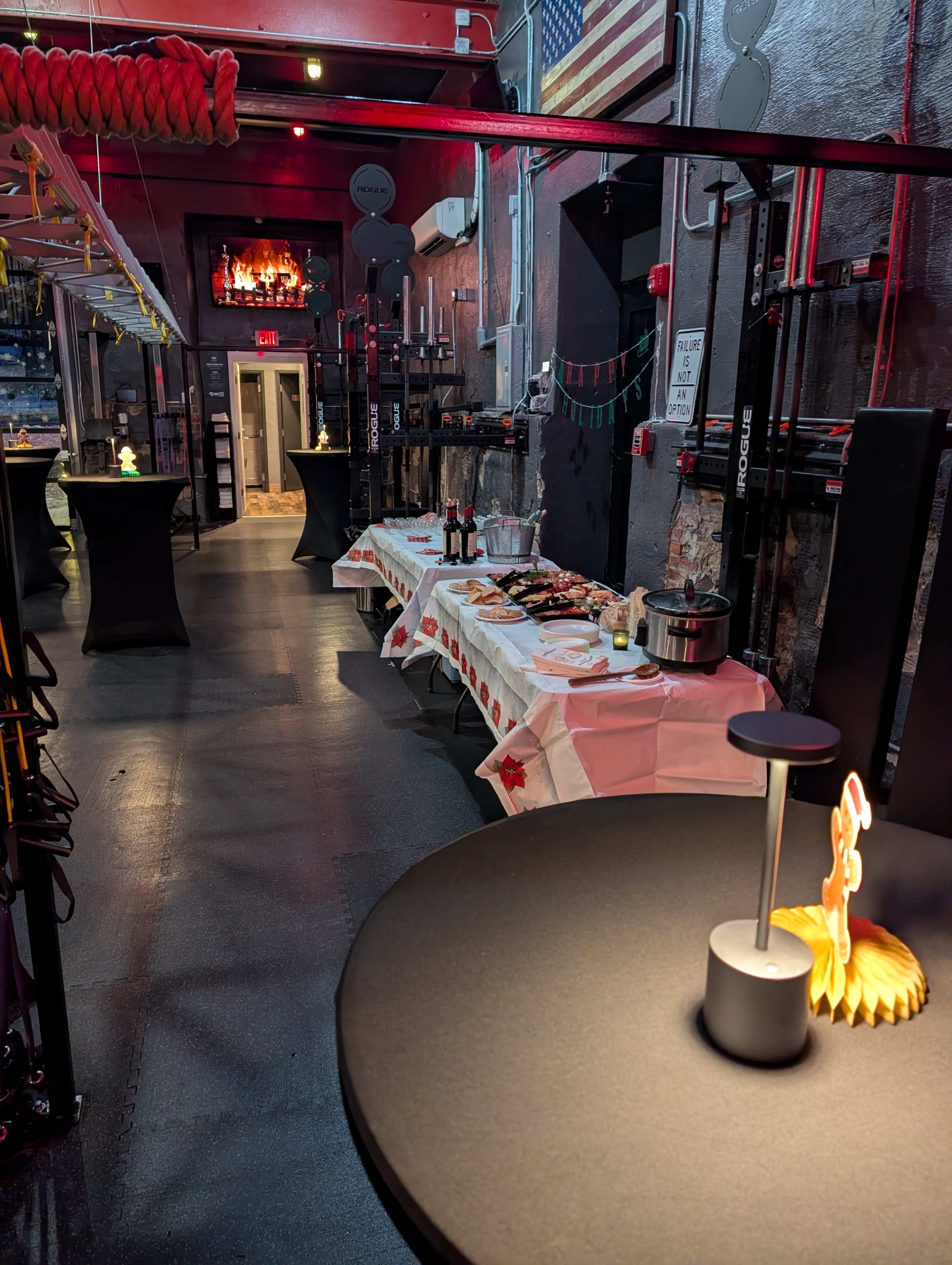 The image shows a gym interior set up for an event, featuring tables with food and drinks alongside black high-top tables.