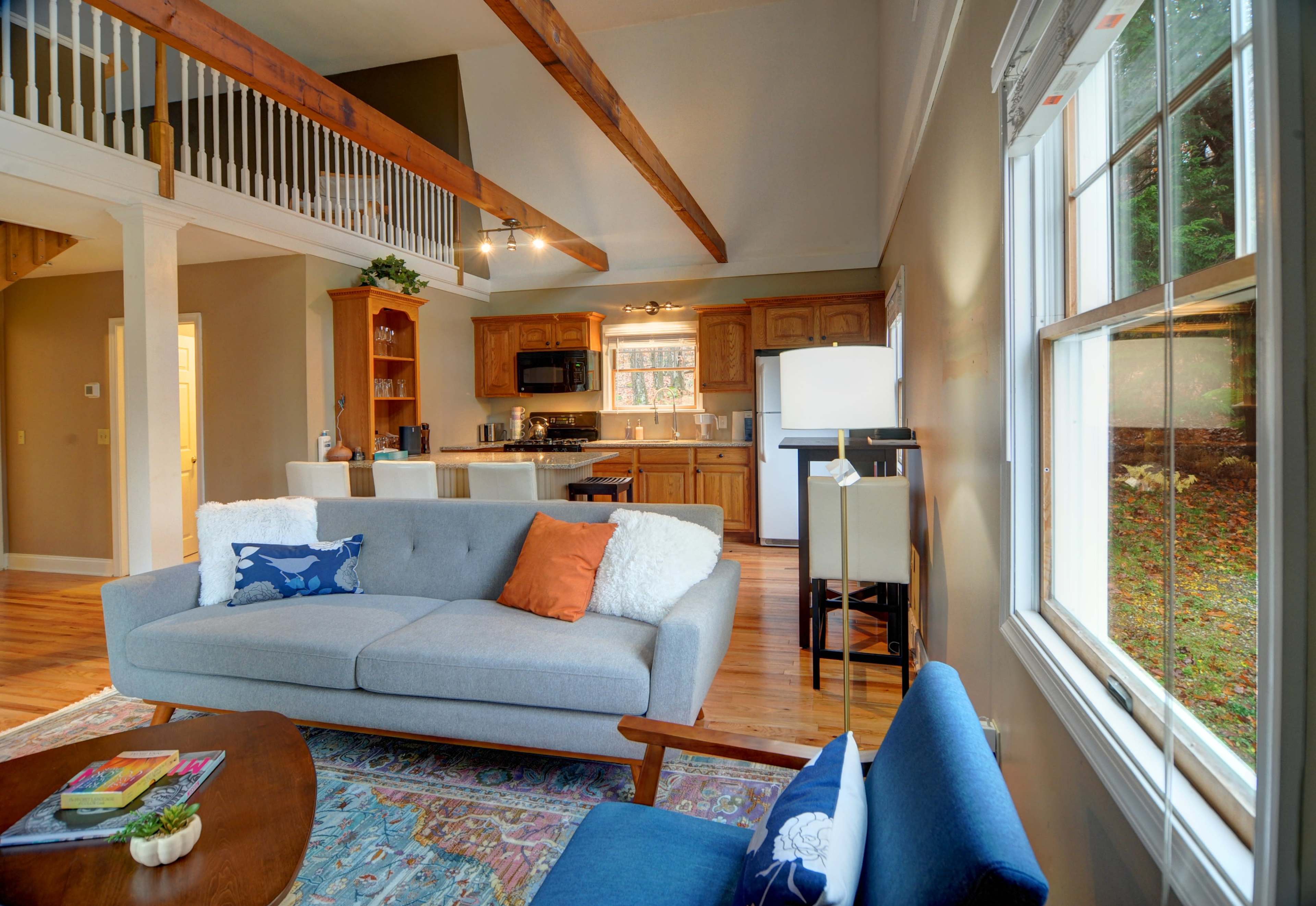 A cozy living room connected to a kitchen, featuring a grey sofa, a wooden coffee table, and a spacious layout with wooden beams and a large window overlooking an outdoor area.