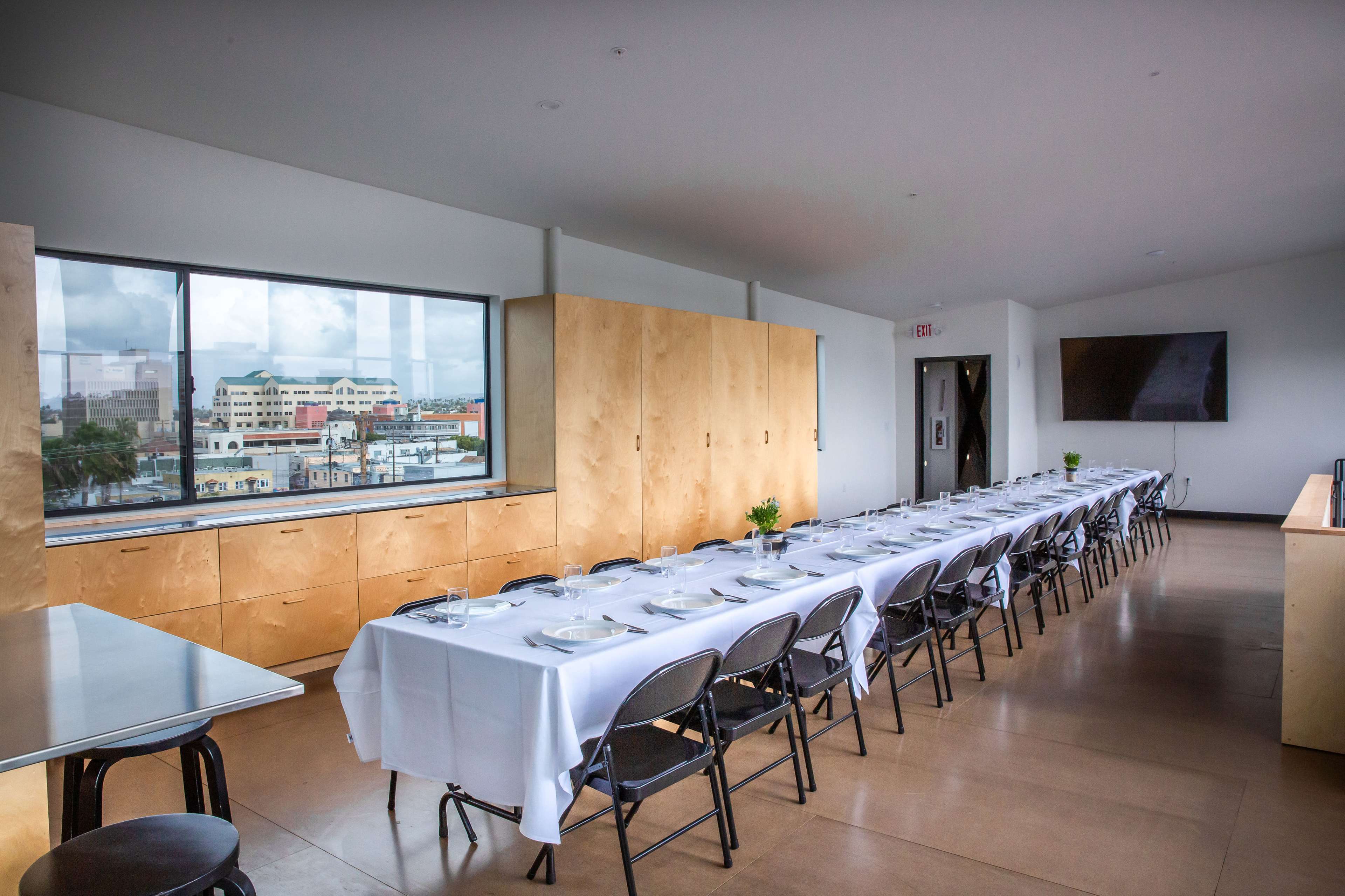 A long table set with white tablecloths and dishware is arranged in a room overlooking a cityscape through large windows.