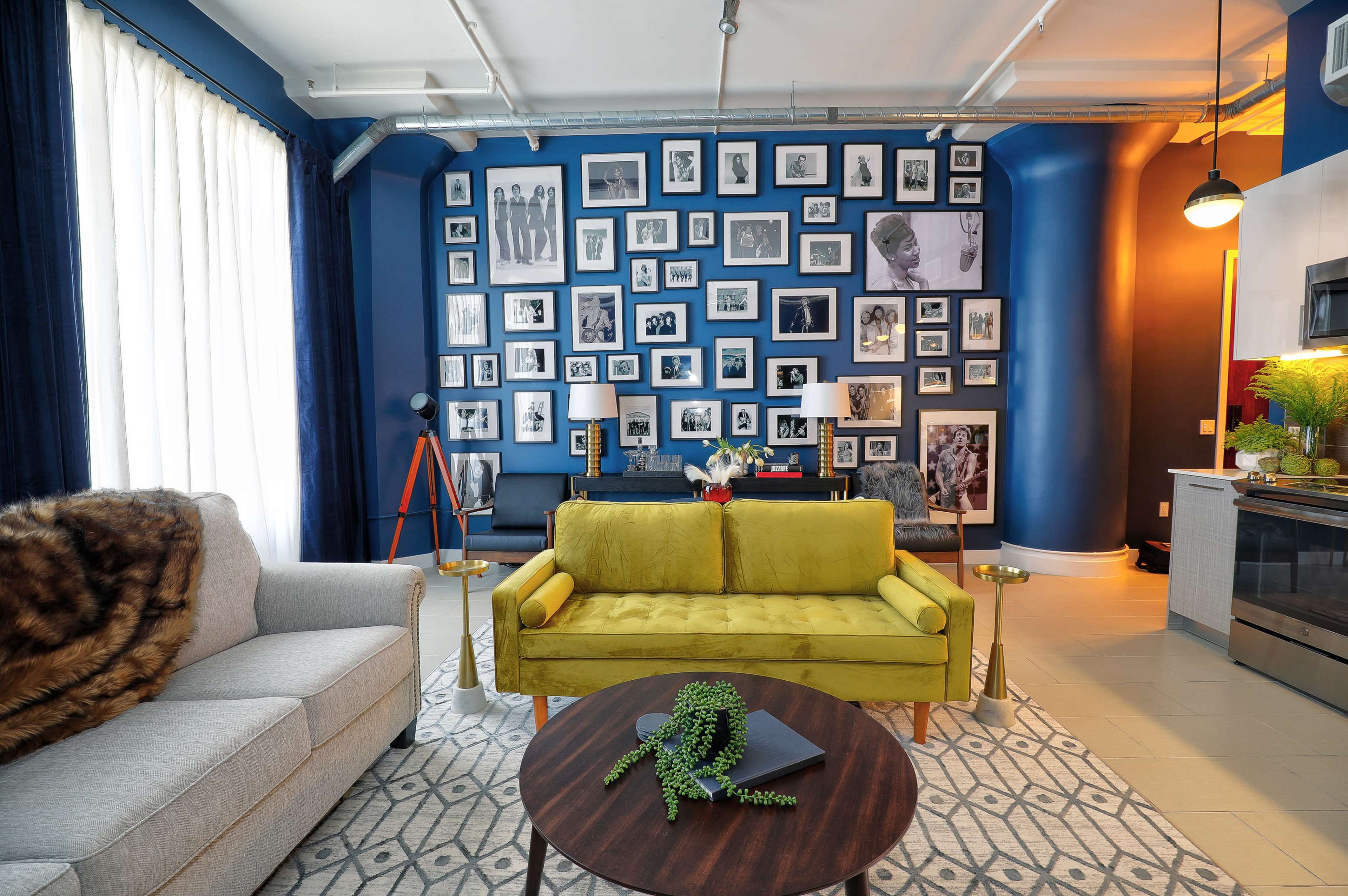 The image shows a living room featuring a yellow sofa, a gray armchair, a round coffee table, and a gallery wall filled with black-and-white photographs.