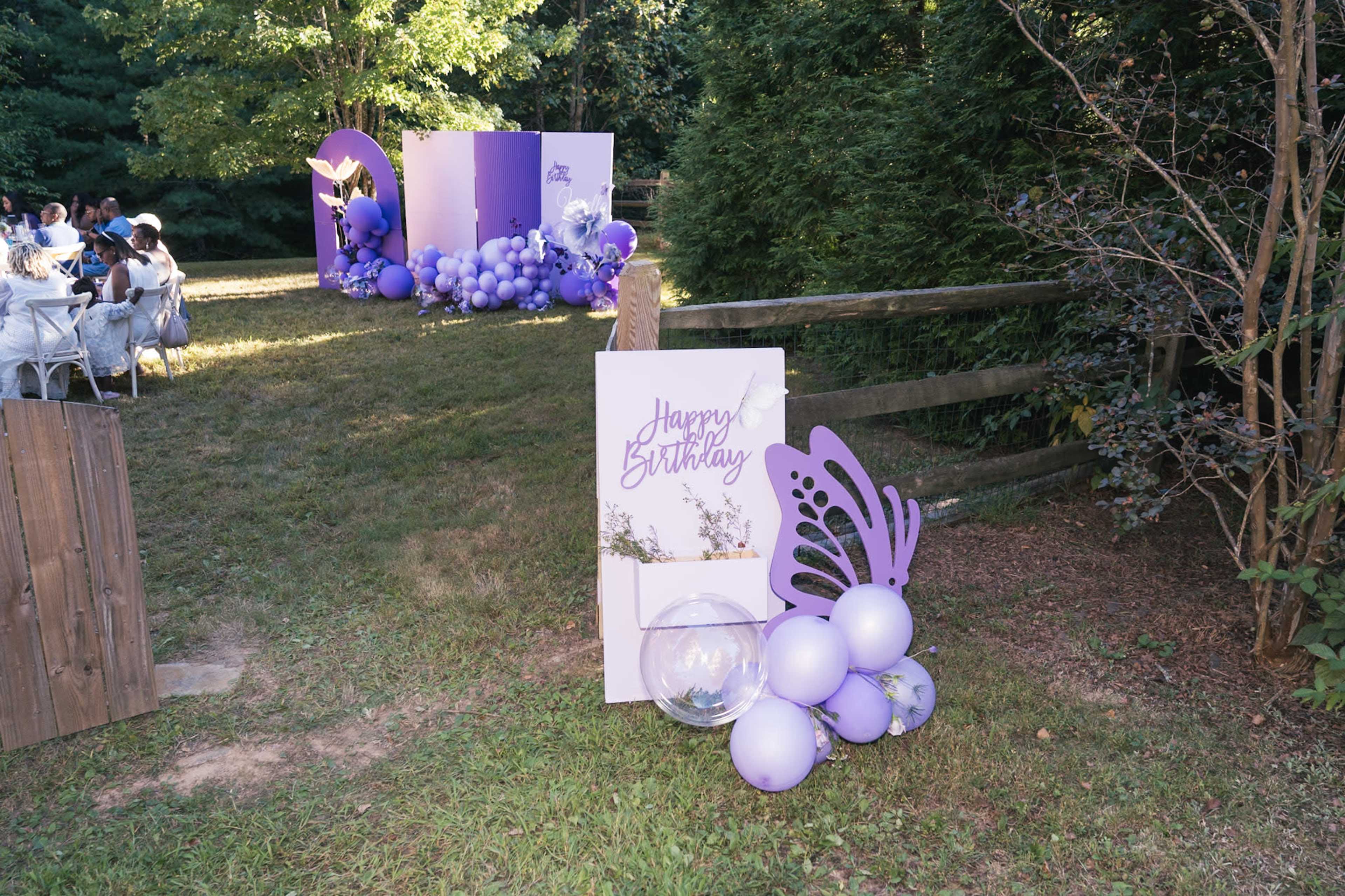 A birthday celebration setup features a "Happy Birthday" sign with purple balloons and a butterfly decoration in a grassy outdoor area.