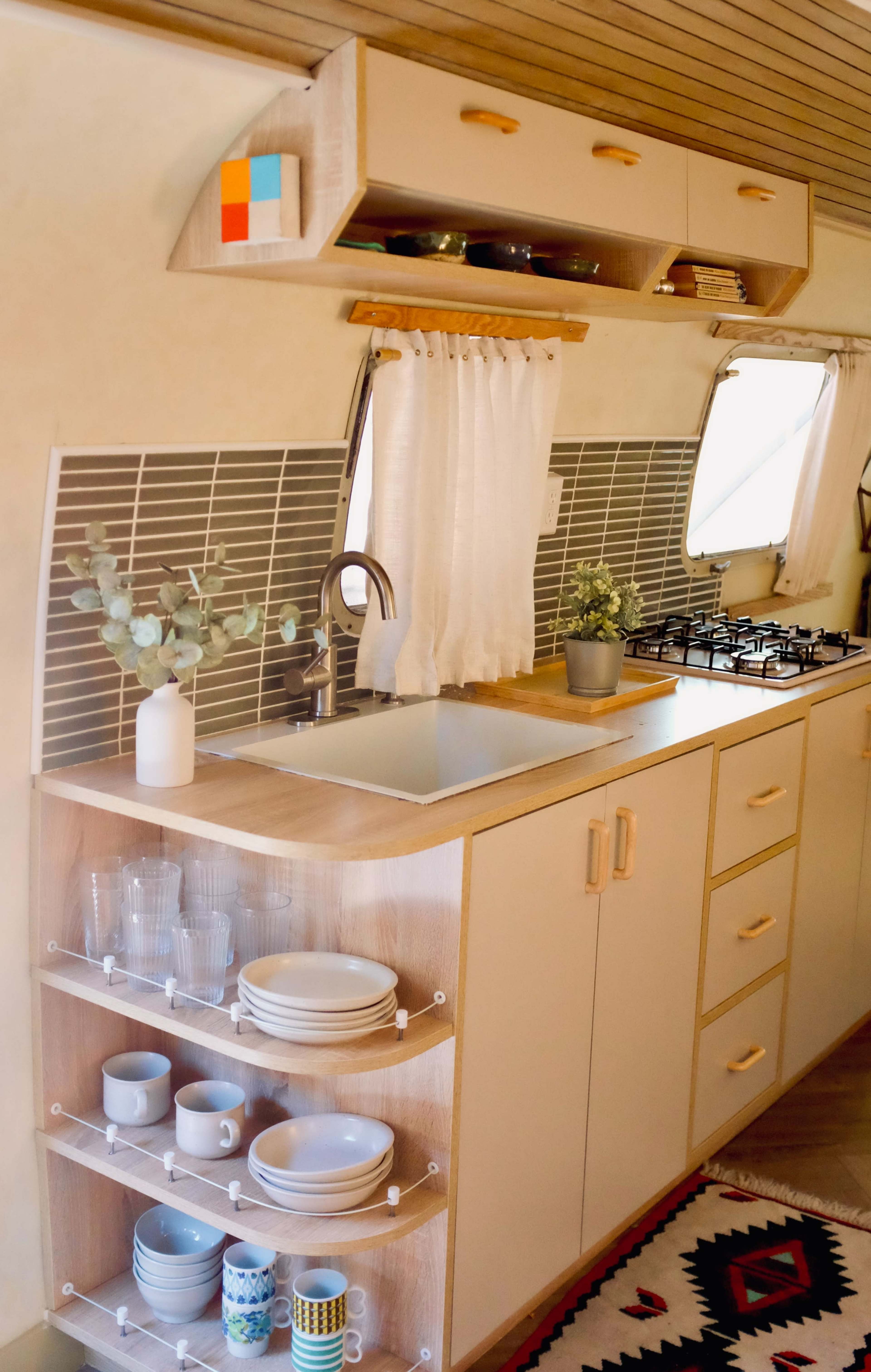 The image shows a compact kitchen area in an RV with wooden cabinetry, a sink, a gas stove, and shelves displaying glassware and dishes.