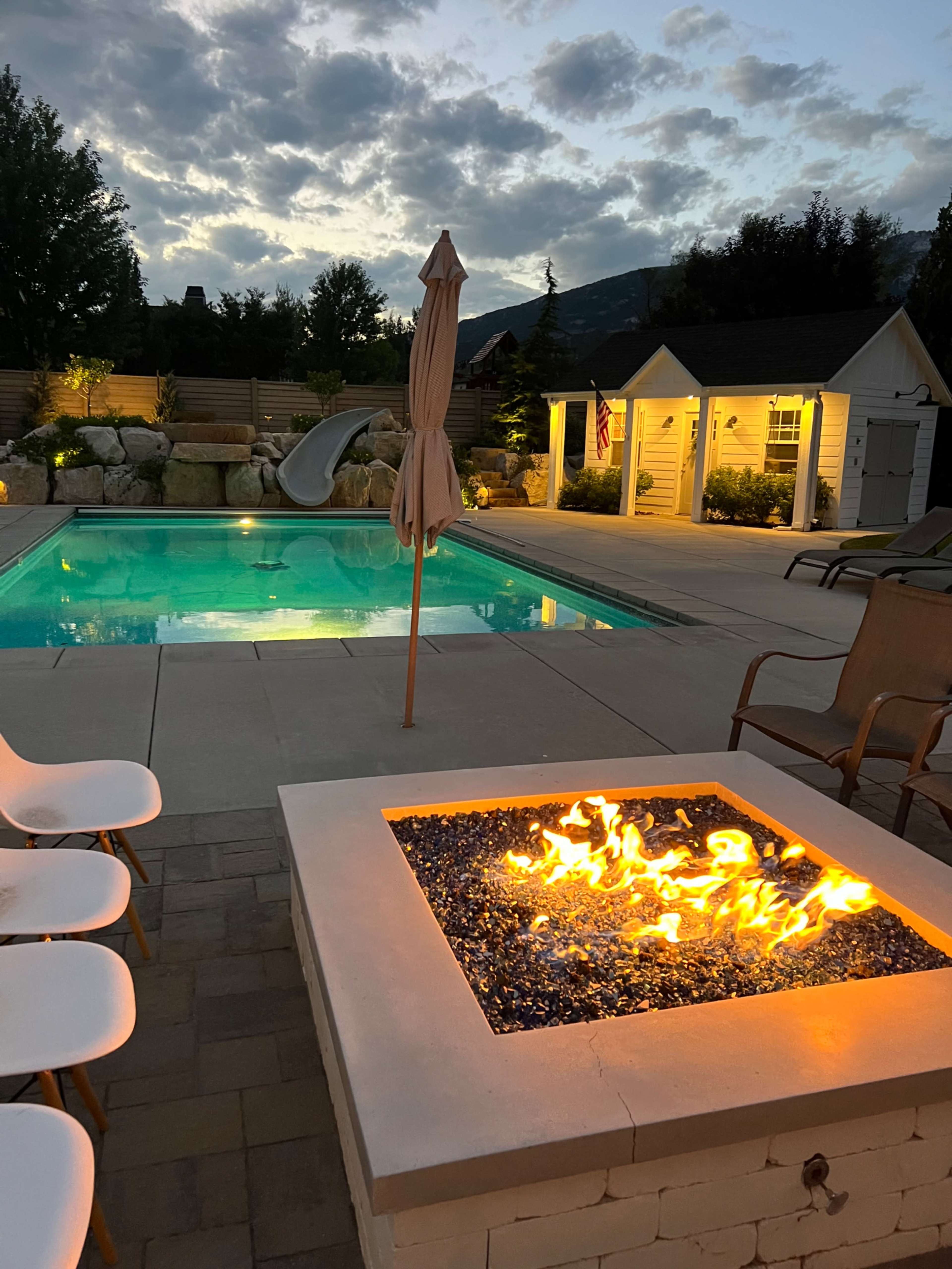 A backyard pool area featuring a lit fire pit in the foreground, a pool with a water slide in the background, and a partially cloudy sky above.