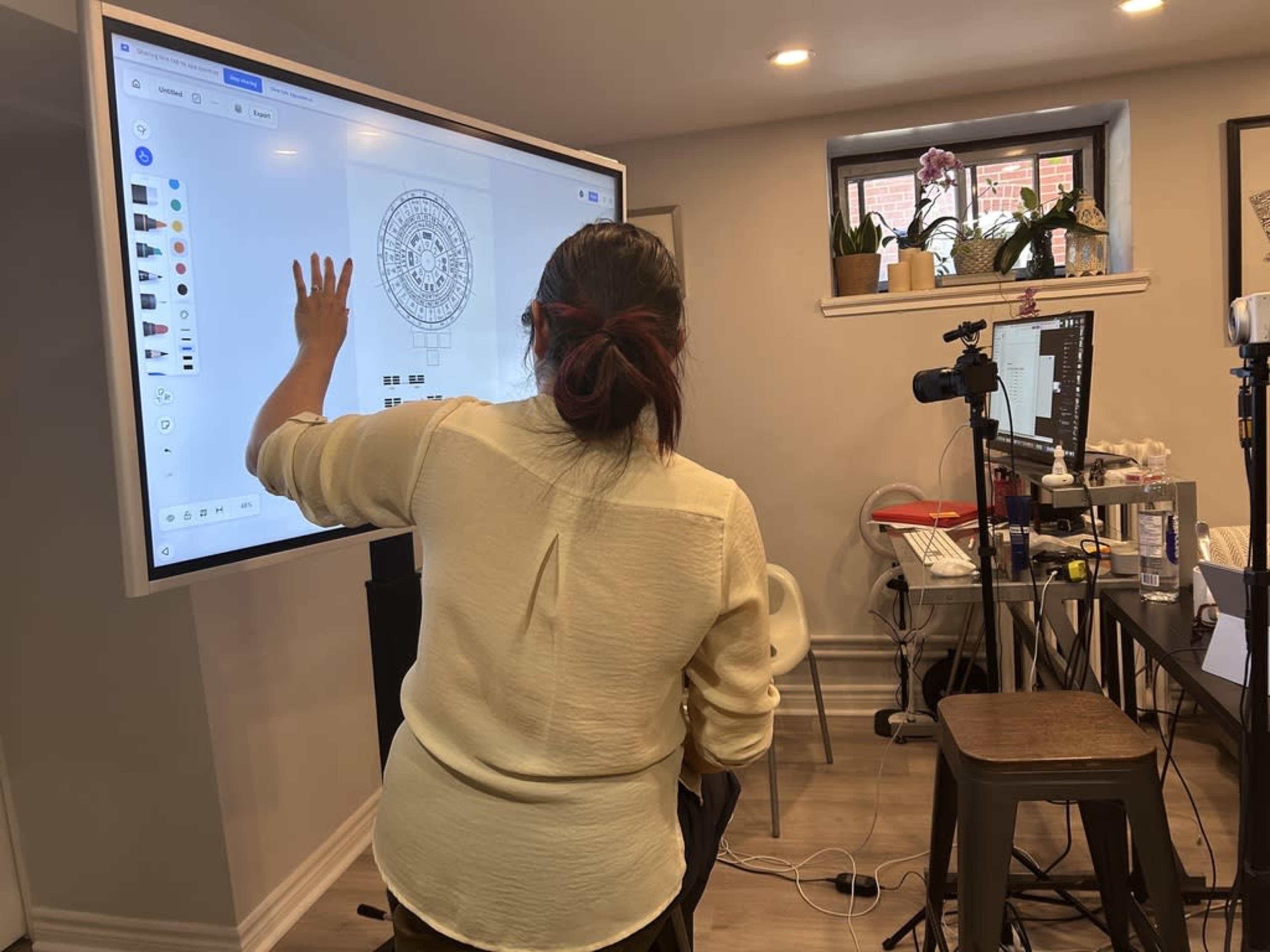 A person is interacting with a large touchscreen display while standing in a well-lit room filled with equipment and furniture.