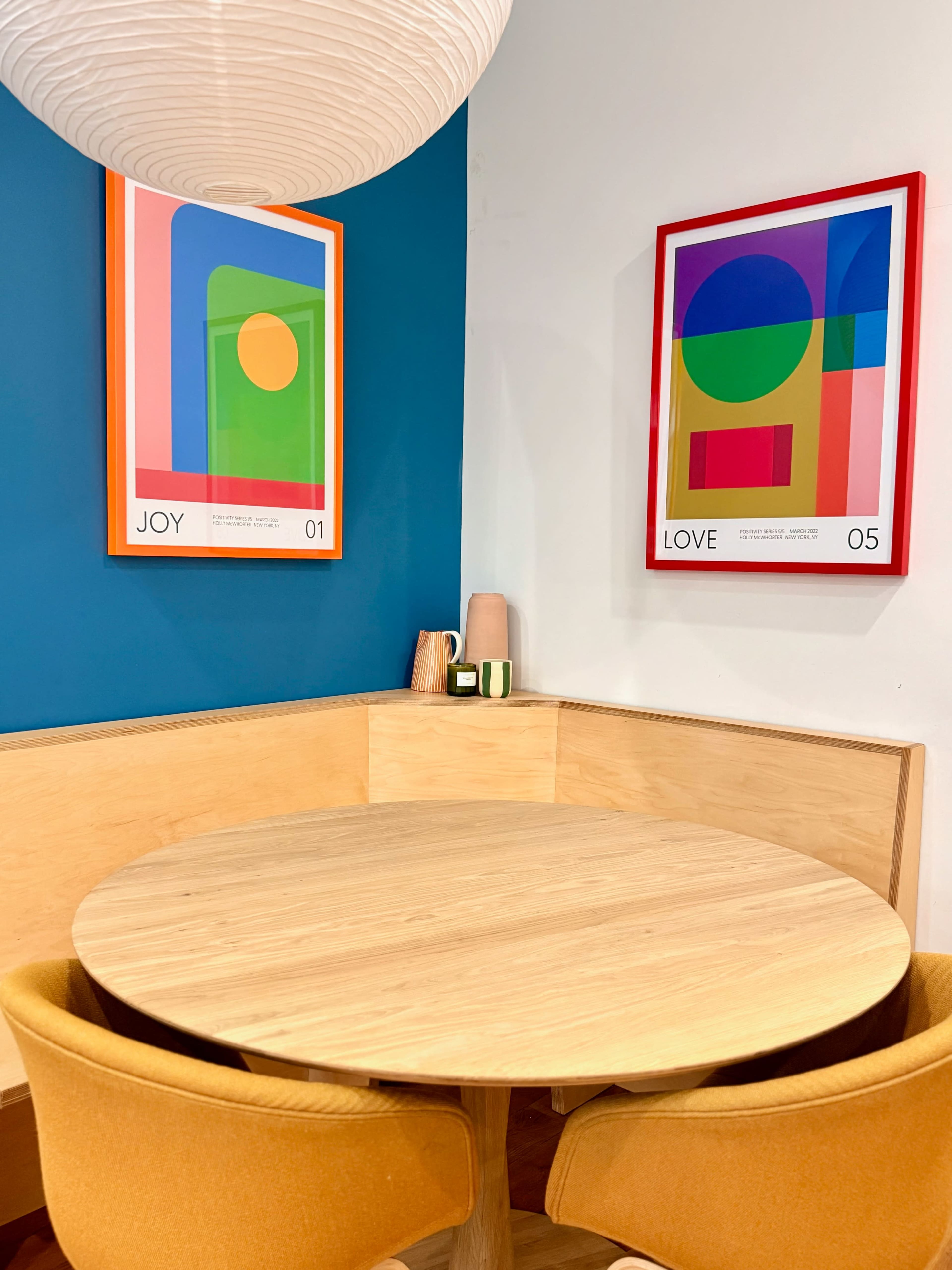 A round wooden table is positioned in a corner of a room, surrounded by orange chairs and decorated with colorful abstract artwork on the walls.