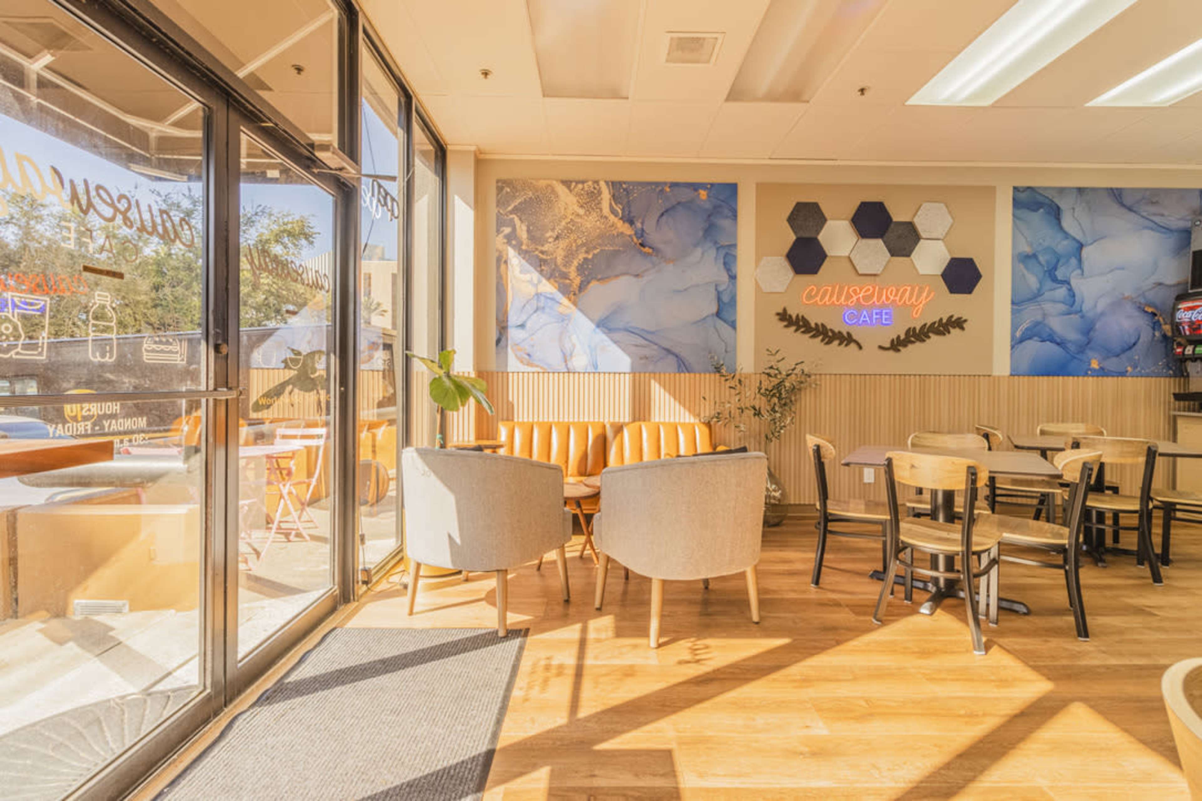 The image shows a cozy café interior with a seating area featuring light-colored chairs and a wooden floor, along with large windows allowing natural light to enter.