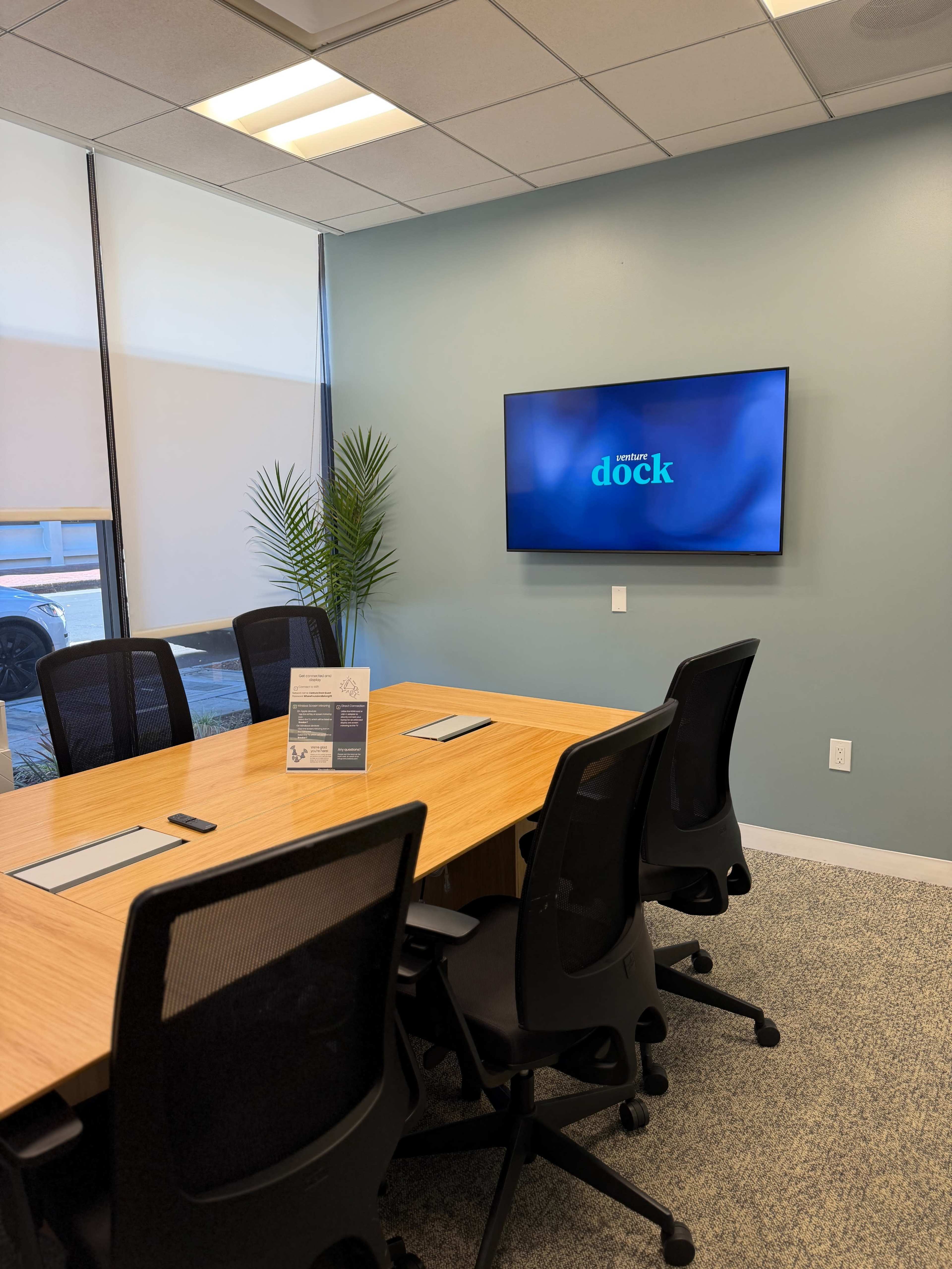 The image shows a modern conference room featuring a wooden meeting table surrounded by black office chairs and a wall-mounted screen displaying the word "dock."
