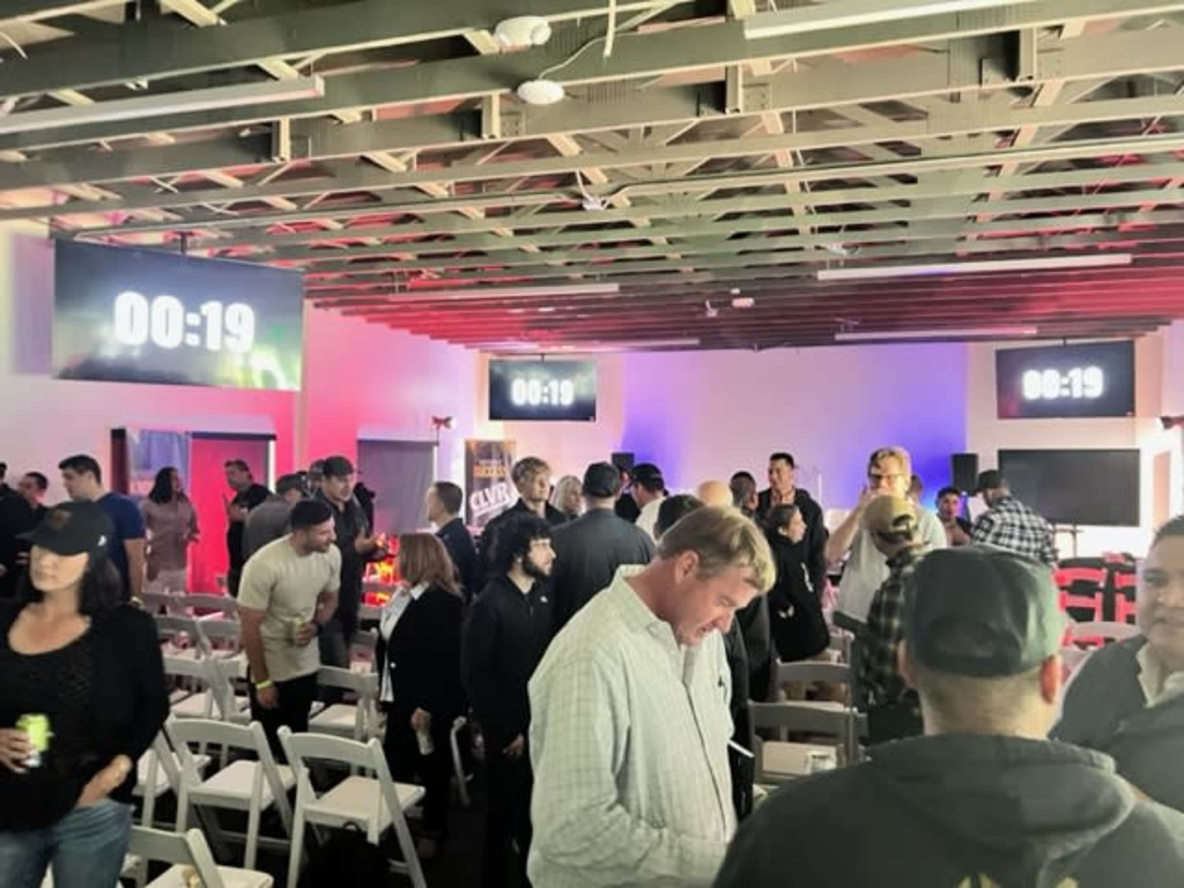 A crowded event space features large digital countdown timers on the walls, with attendees mingling and sitting in white chairs.