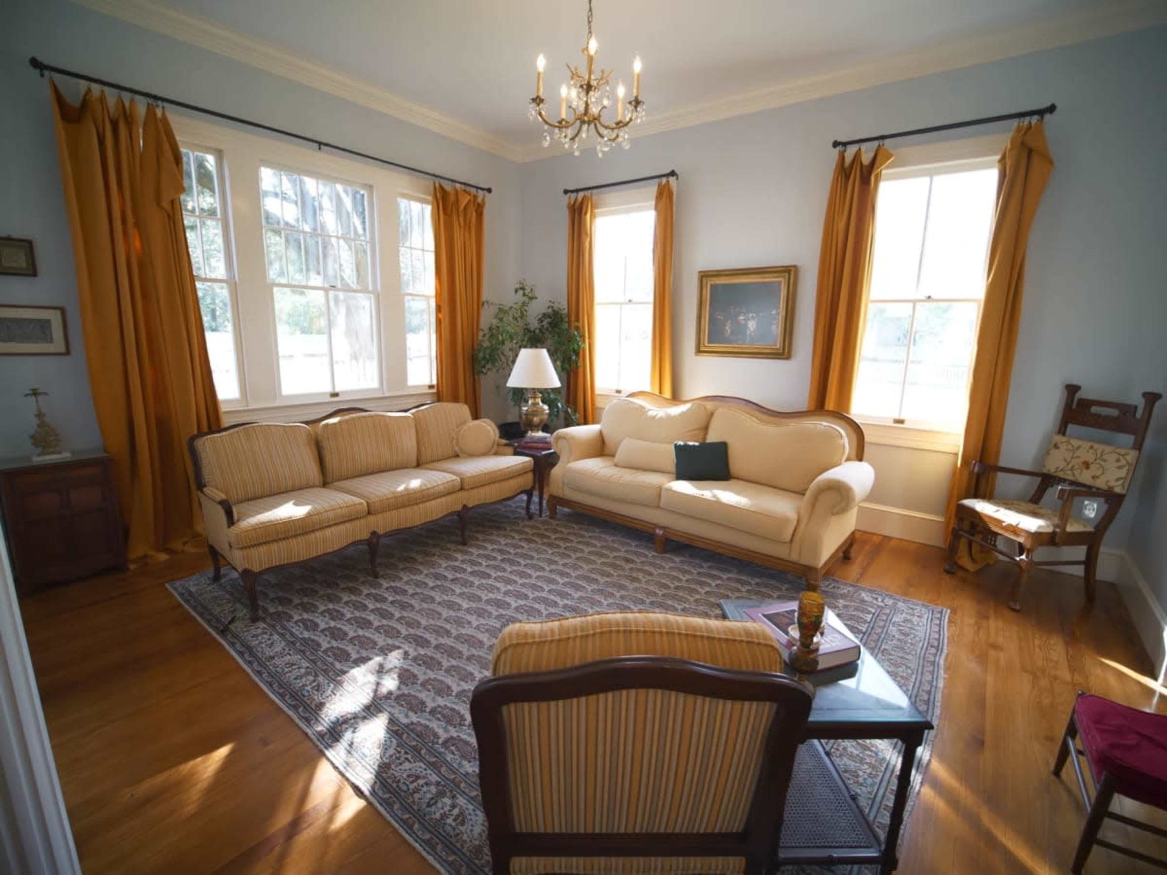 A well-lit living room featuring a cream-colored sofa set, a decorative area rug, and large windows with yellow curtains.