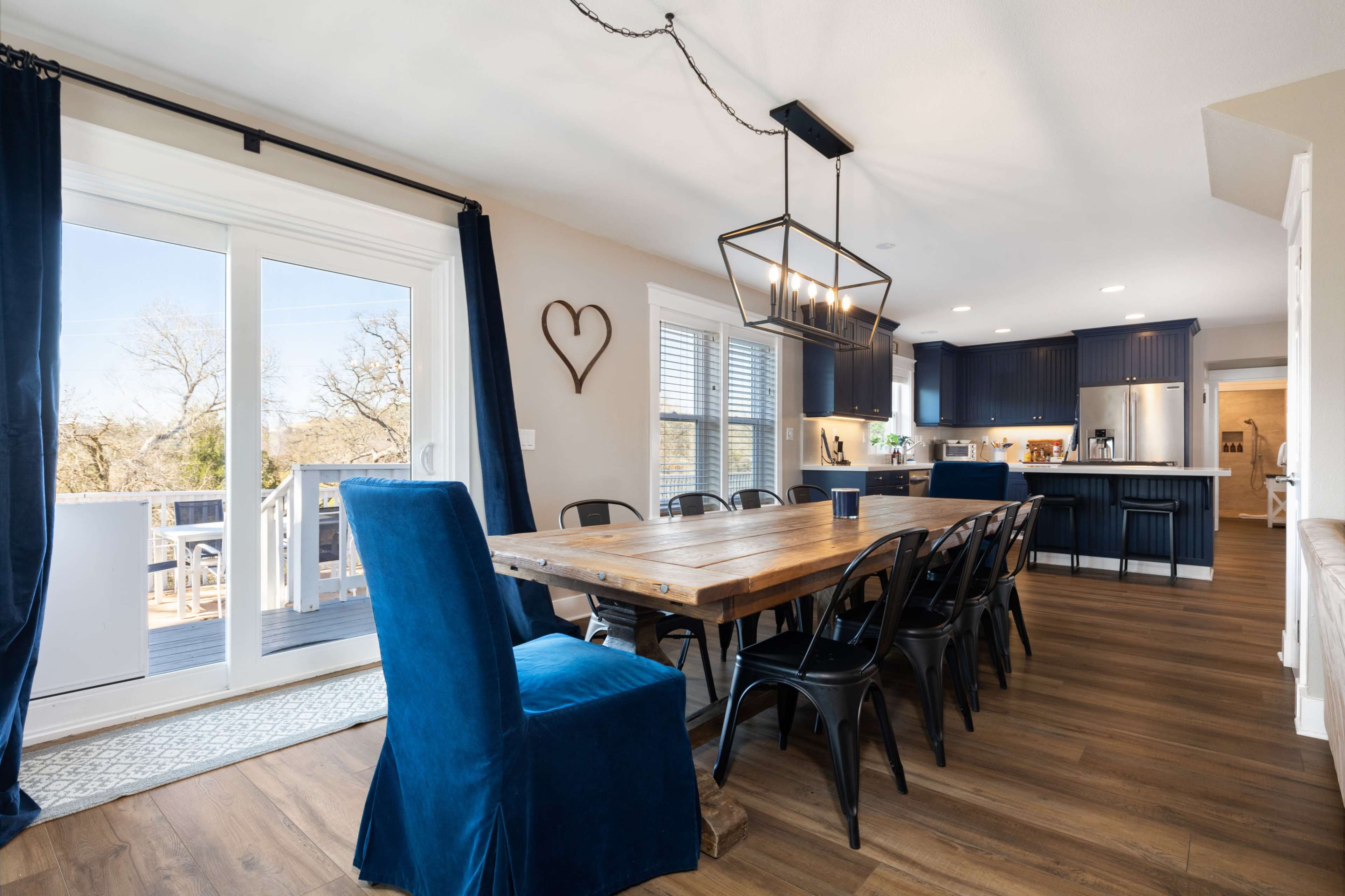 A spacious dining area features a wooden table surrounded by black chairs and blue upholstered chairs, with large windows and sliding doors leading to an outdoor deck.