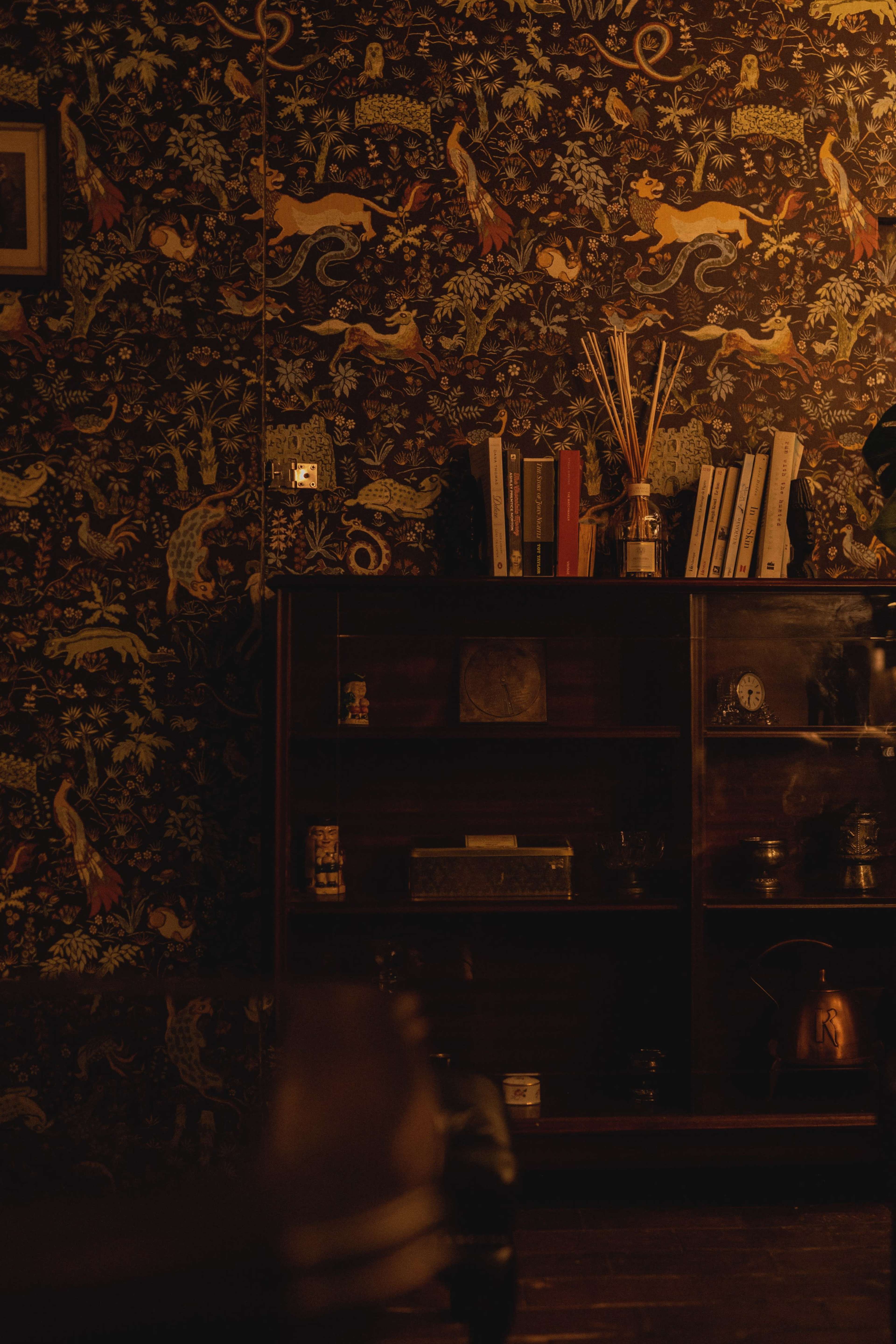 A wooden shelf filled with books and decorative items stands against a patterned wallpaper, illuminated by soft ambient lighting.
