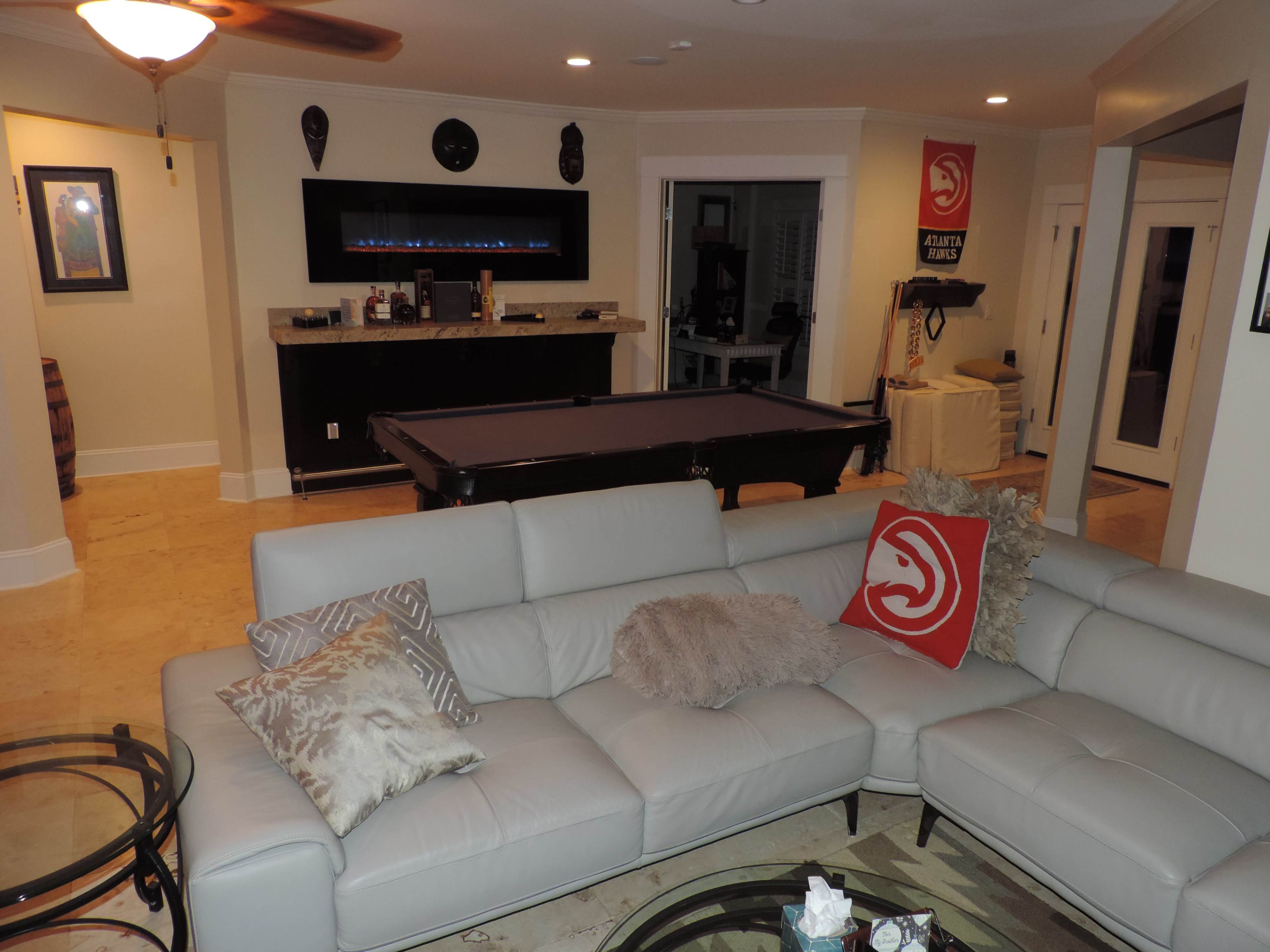 The image shows a living room with a light-colored sectional sofa, a pool table in the background, and various decorations on the walls.