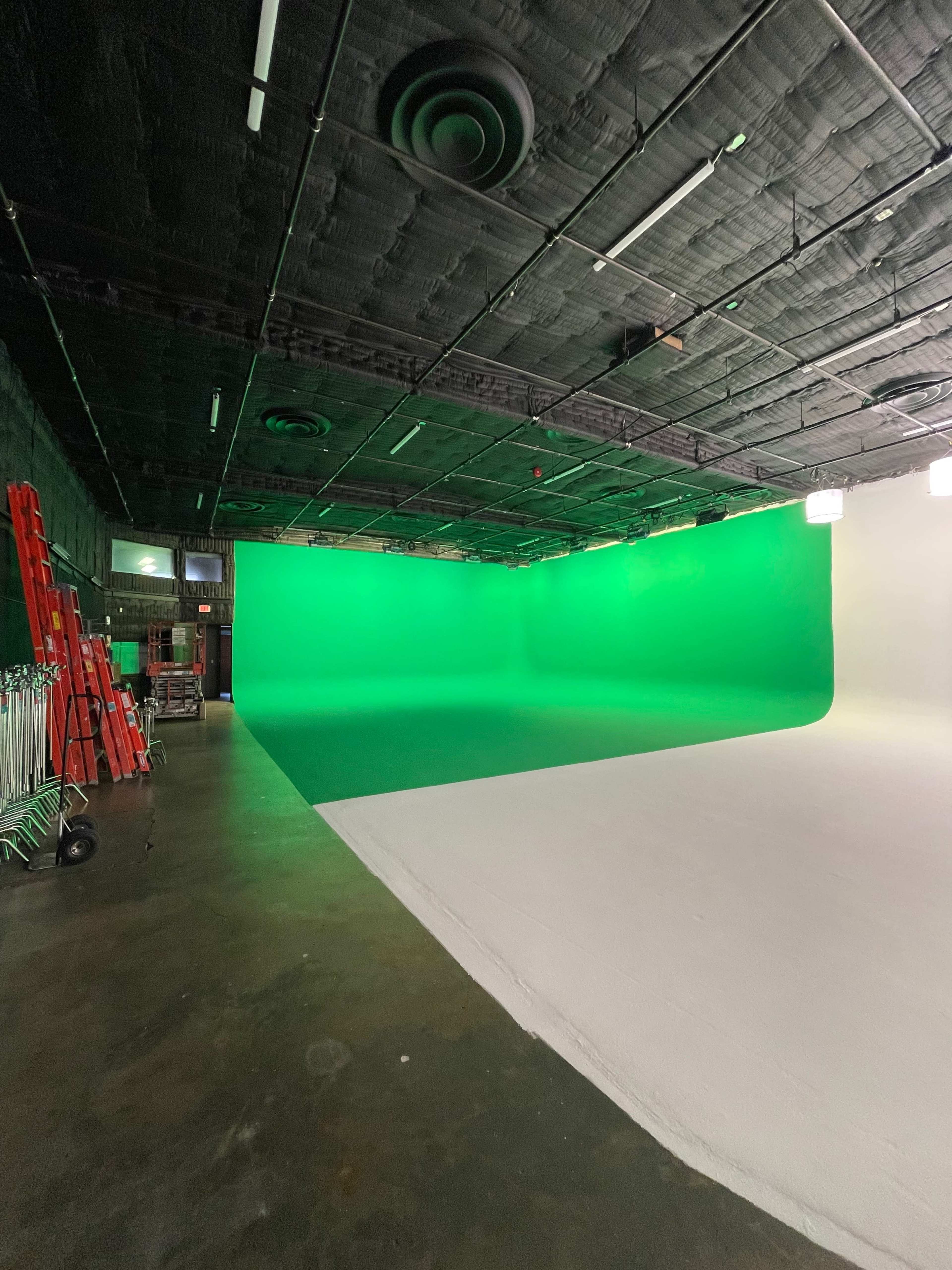 A studio with a green screen on one side and a white surface on the other, surrounded by lighting equipment and ladders.
