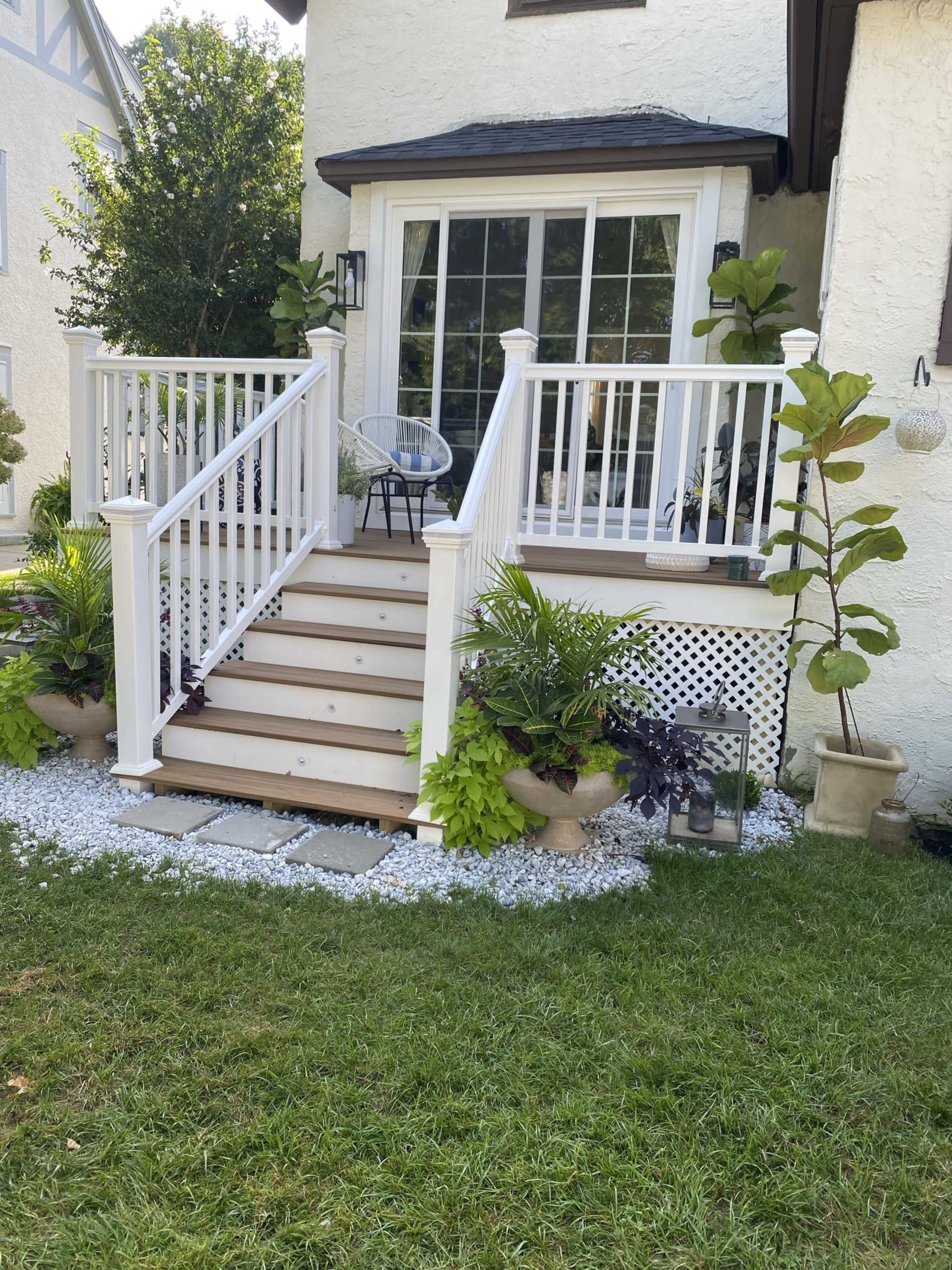 The image shows a set of wooden stairs leading up to a house with a white railing, surrounded by potted plants and decorative stones on the lawn.