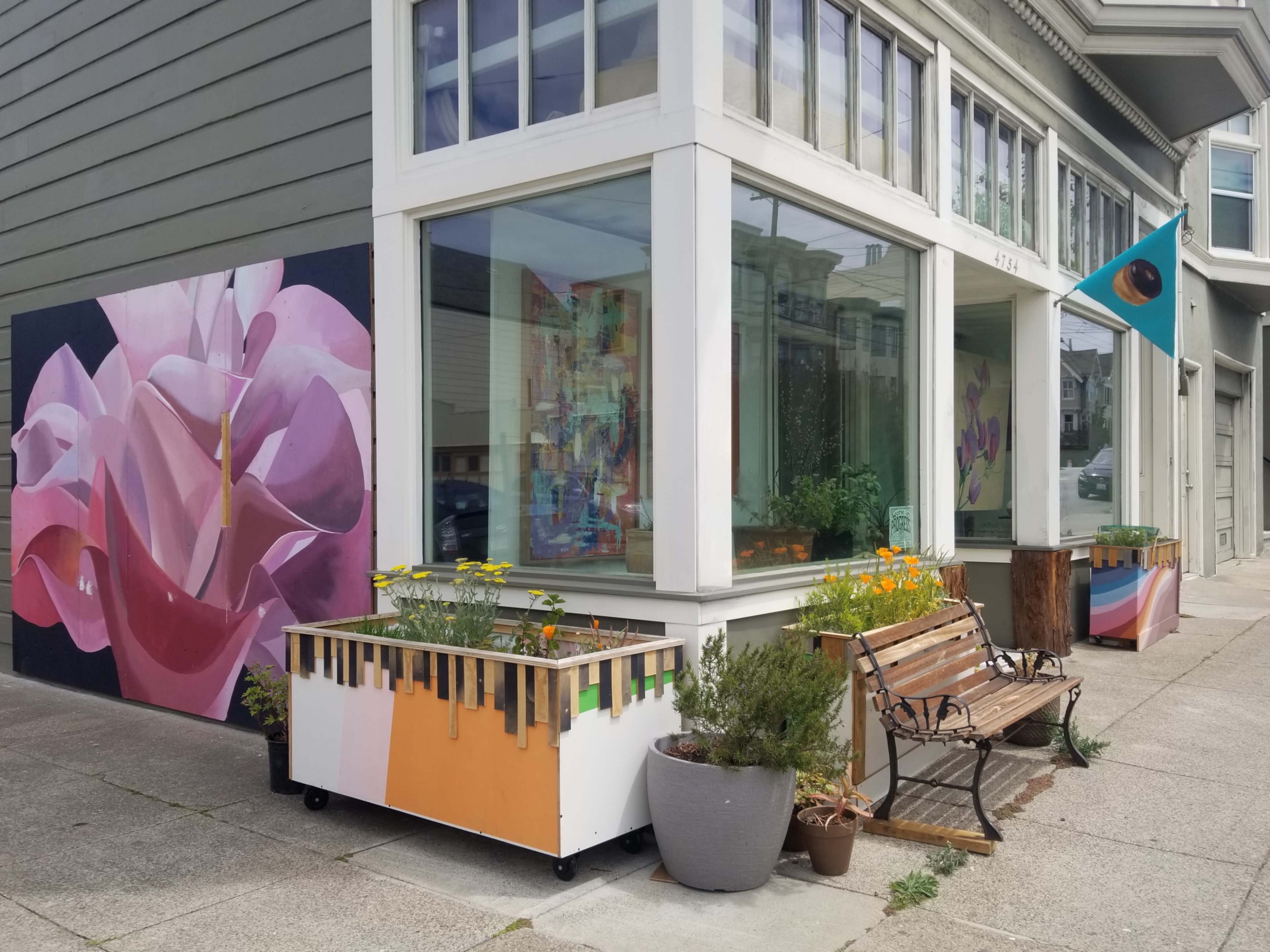 A colorful mural of a pink flower decorates the side of a building, which features large windows, planters with flowers, and a bench in front.