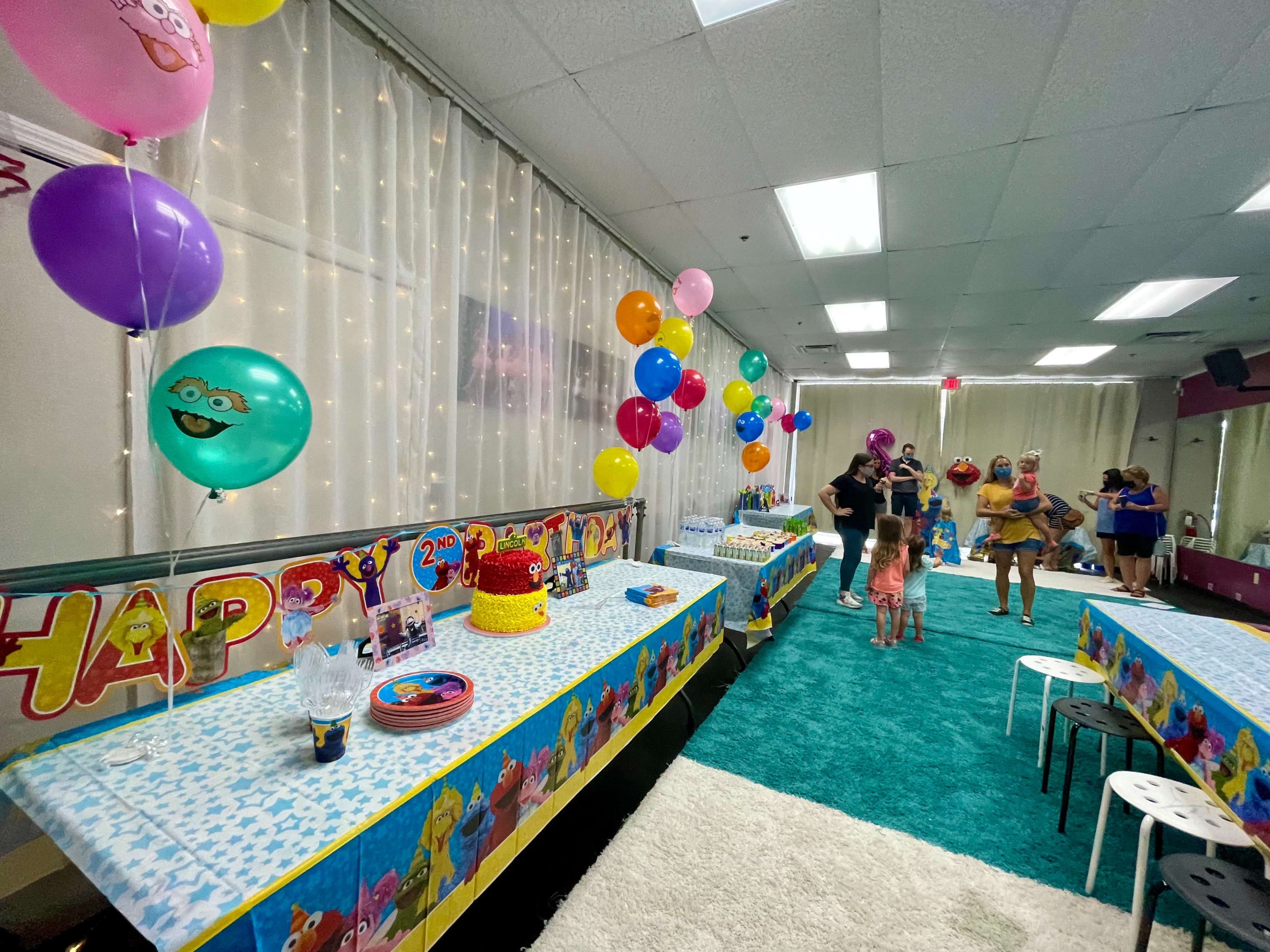 A colorful children's birthday party setup features decorated tables, balloons, and a cake, with guests mingling in a brightly lit room.