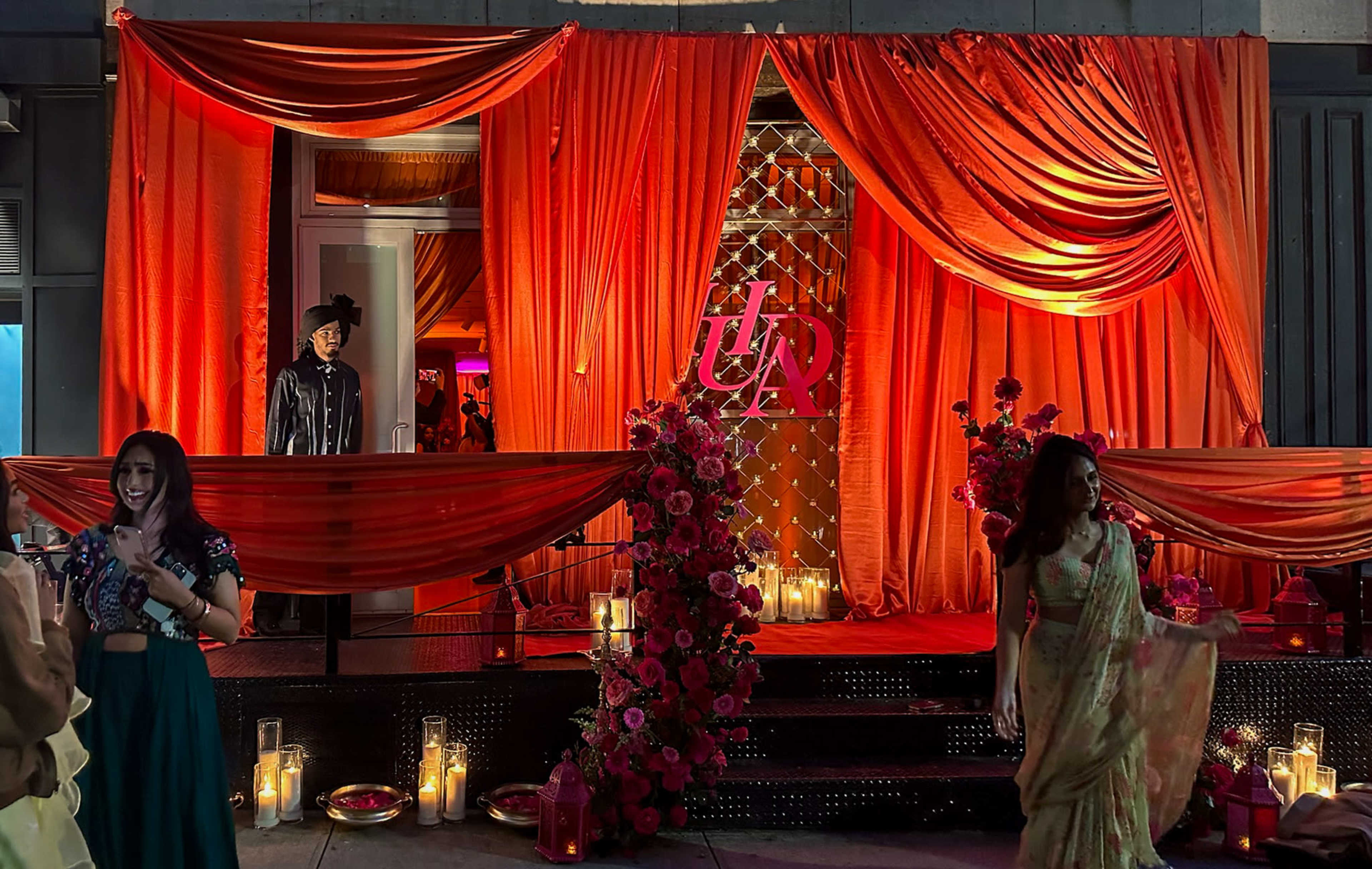 The scene features a brightly decorated entrance adorned with red drapery and floral arrangements, with people mingling outside.