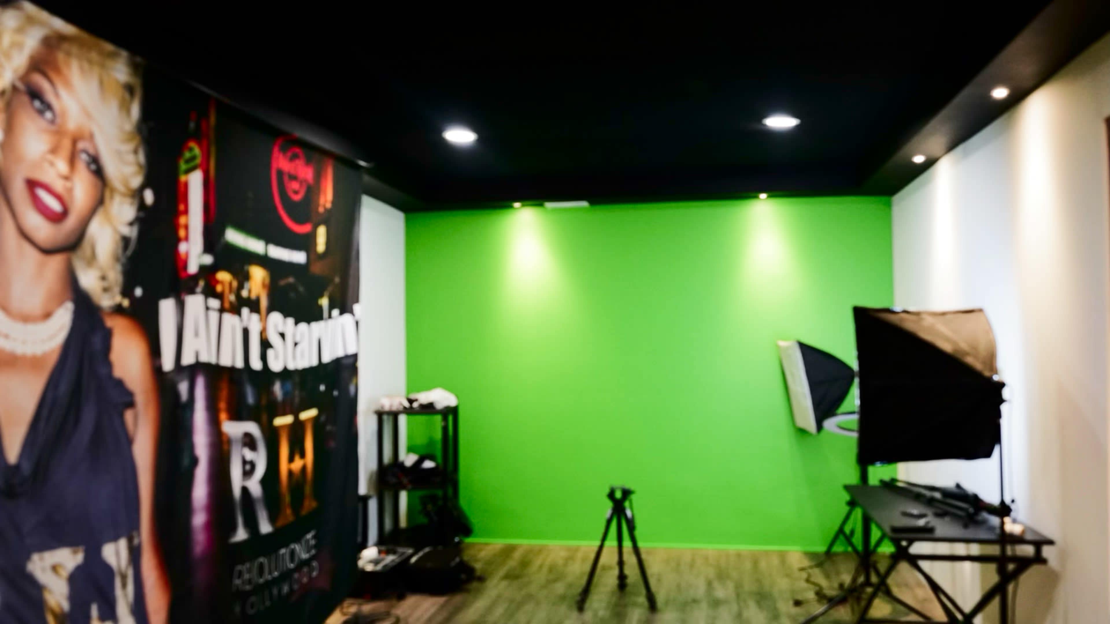 An empty studio space featuring a green backdrop, a camera tripod, and lighting equipment against a wall with a large mural.
