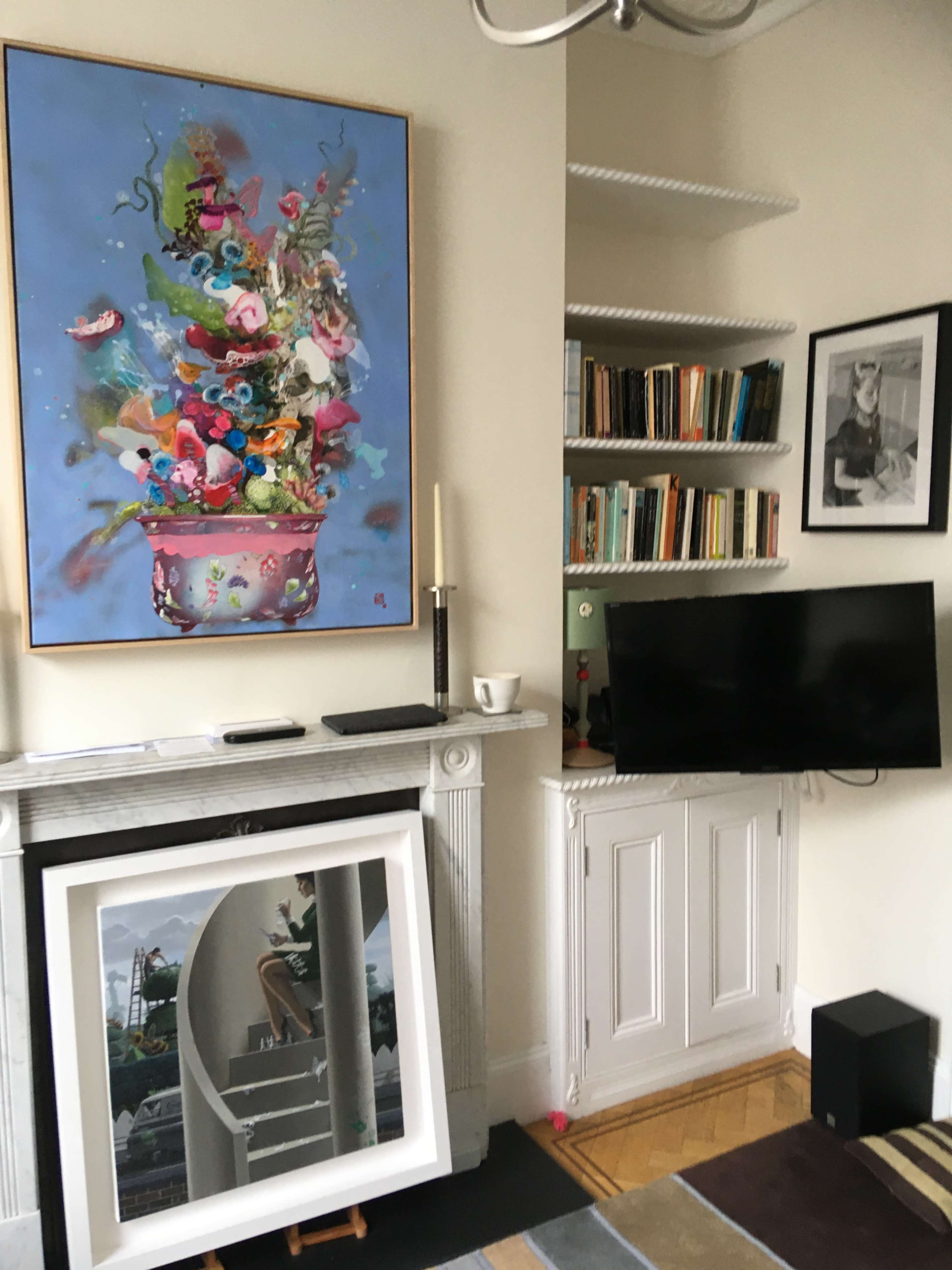 A living room with a colorful painting above a mantel, a shelf of books, a flat-screen TV, and a framed black-and-white photo on the wall.