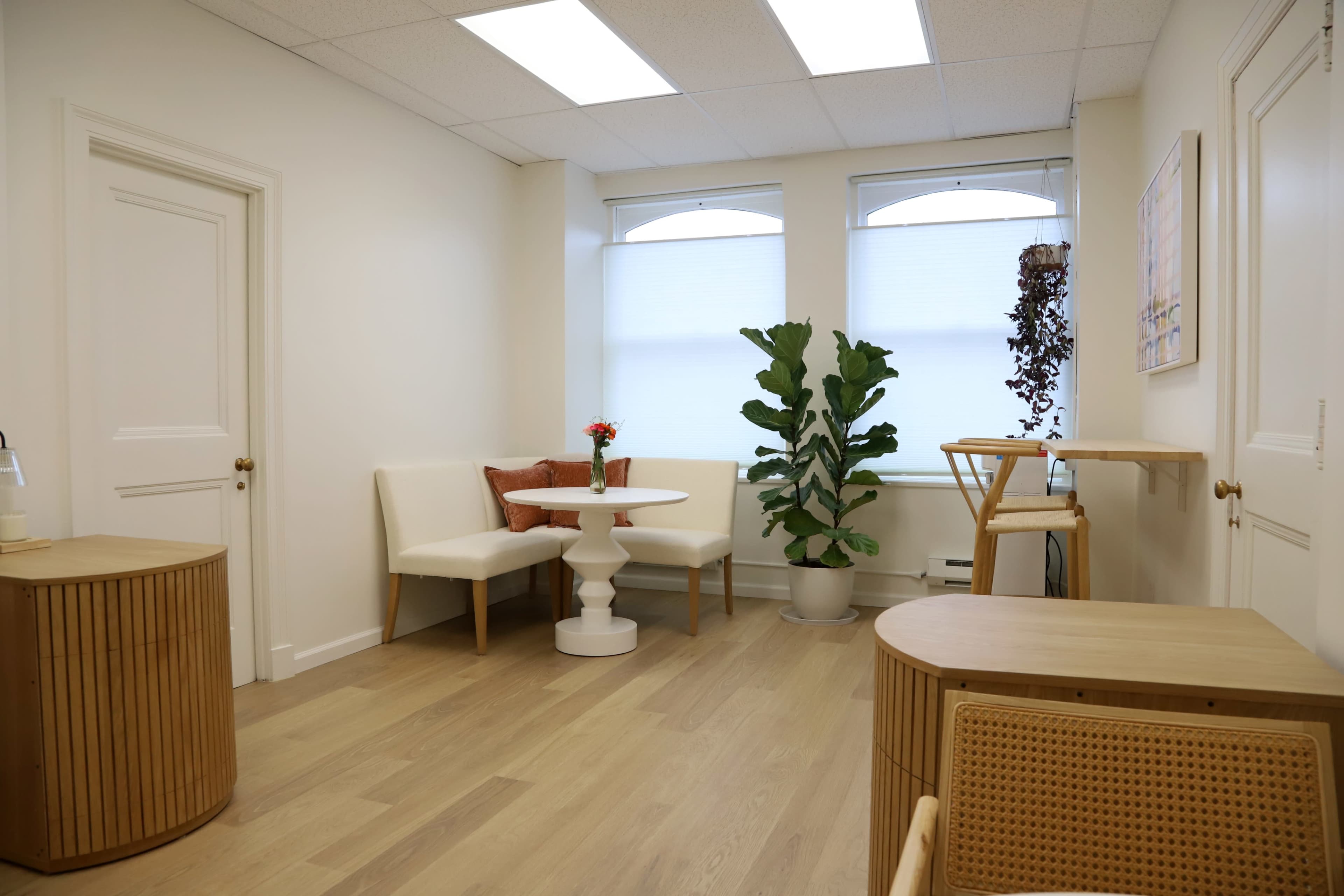 The image shows a bright, minimalistic office space with two seating areas, a plant, and a small table.