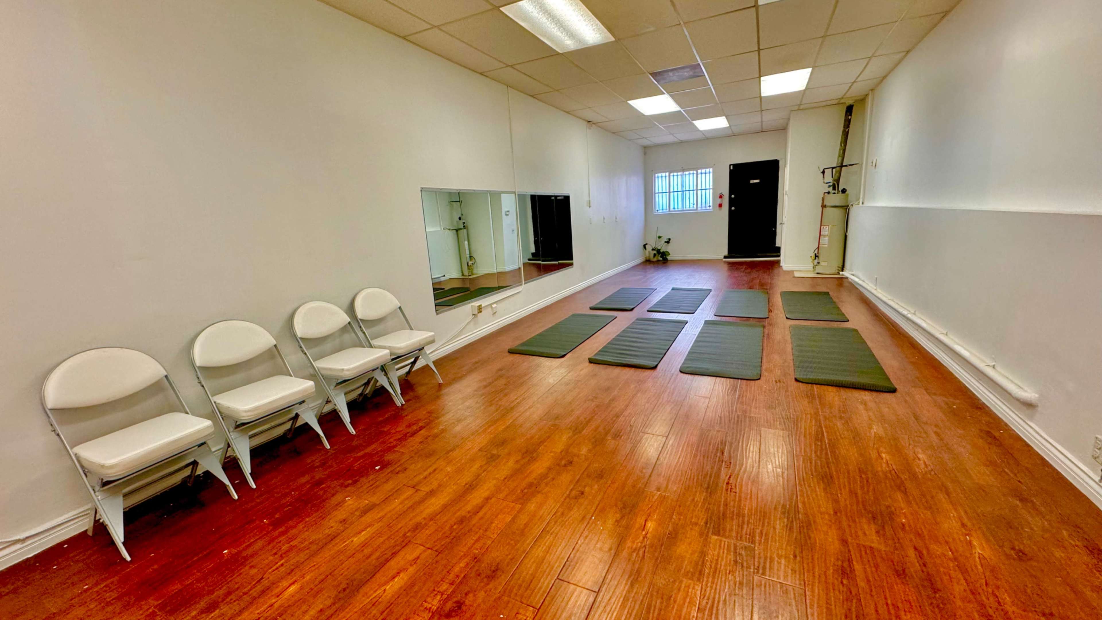 A spacious, well-lit room with wooden flooring, featuring several exercise mats arranged on the floor and a row of folding chairs against one wall.
