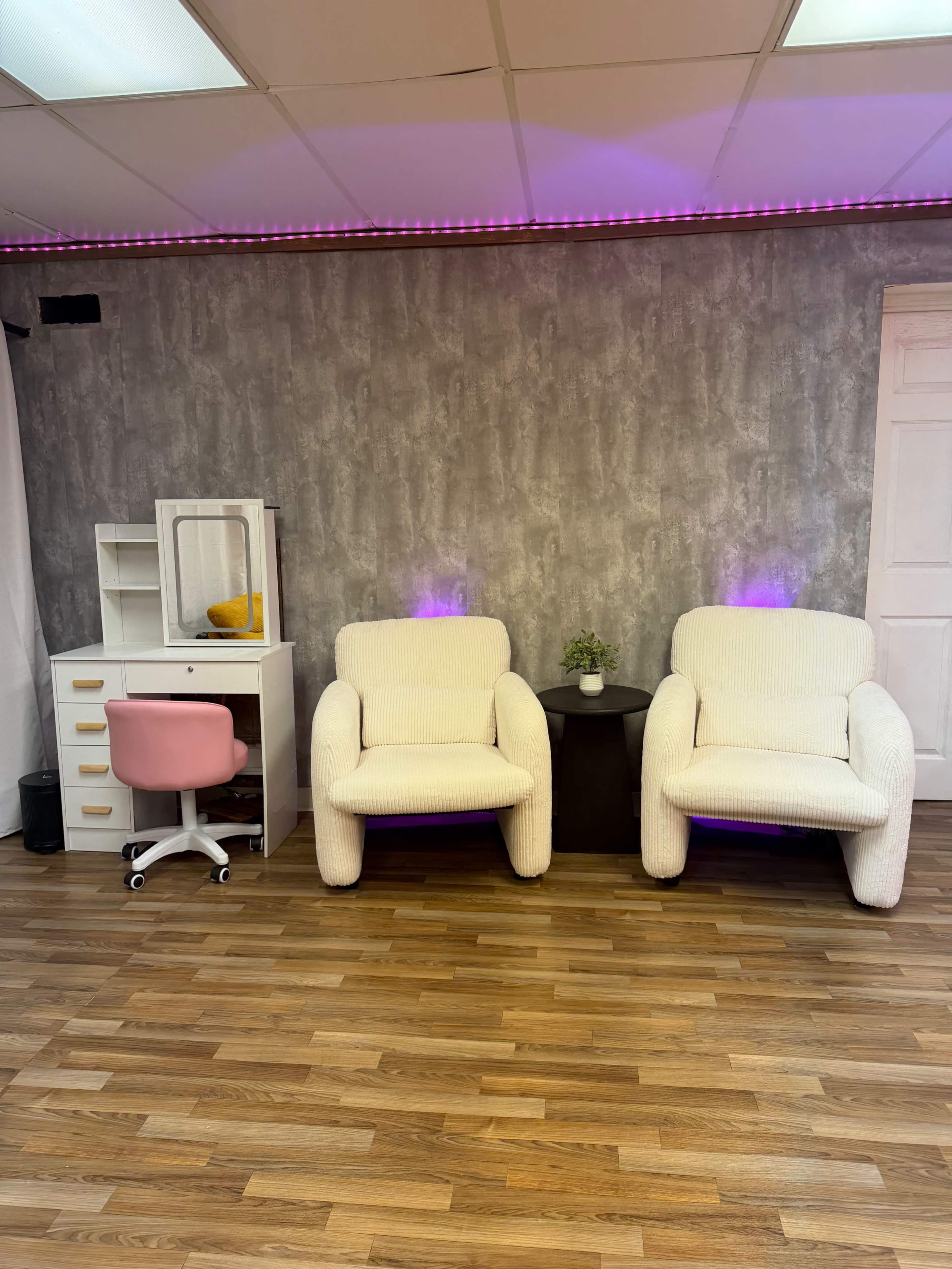 The image shows a room with two textured armchairs, a small side table, and a white desk with a pink chair against a gray wall.