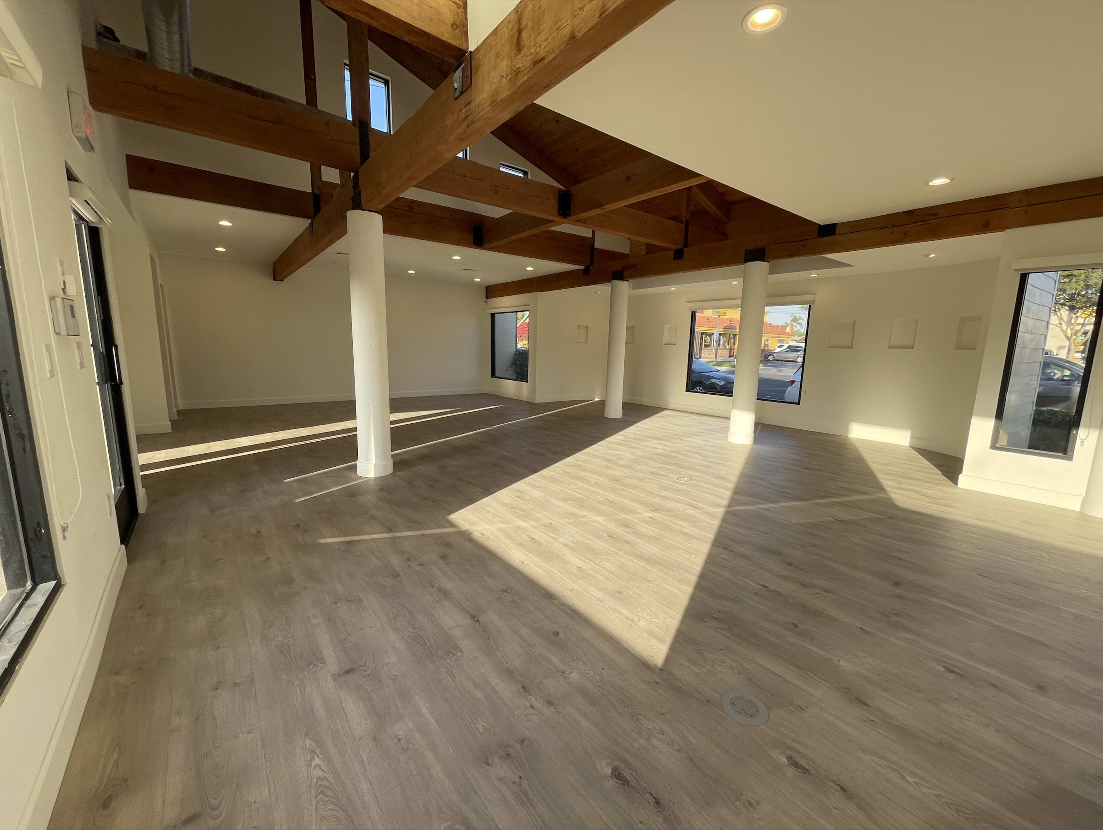 The image shows an empty room with wooden beams, large windows, and laminate flooring.