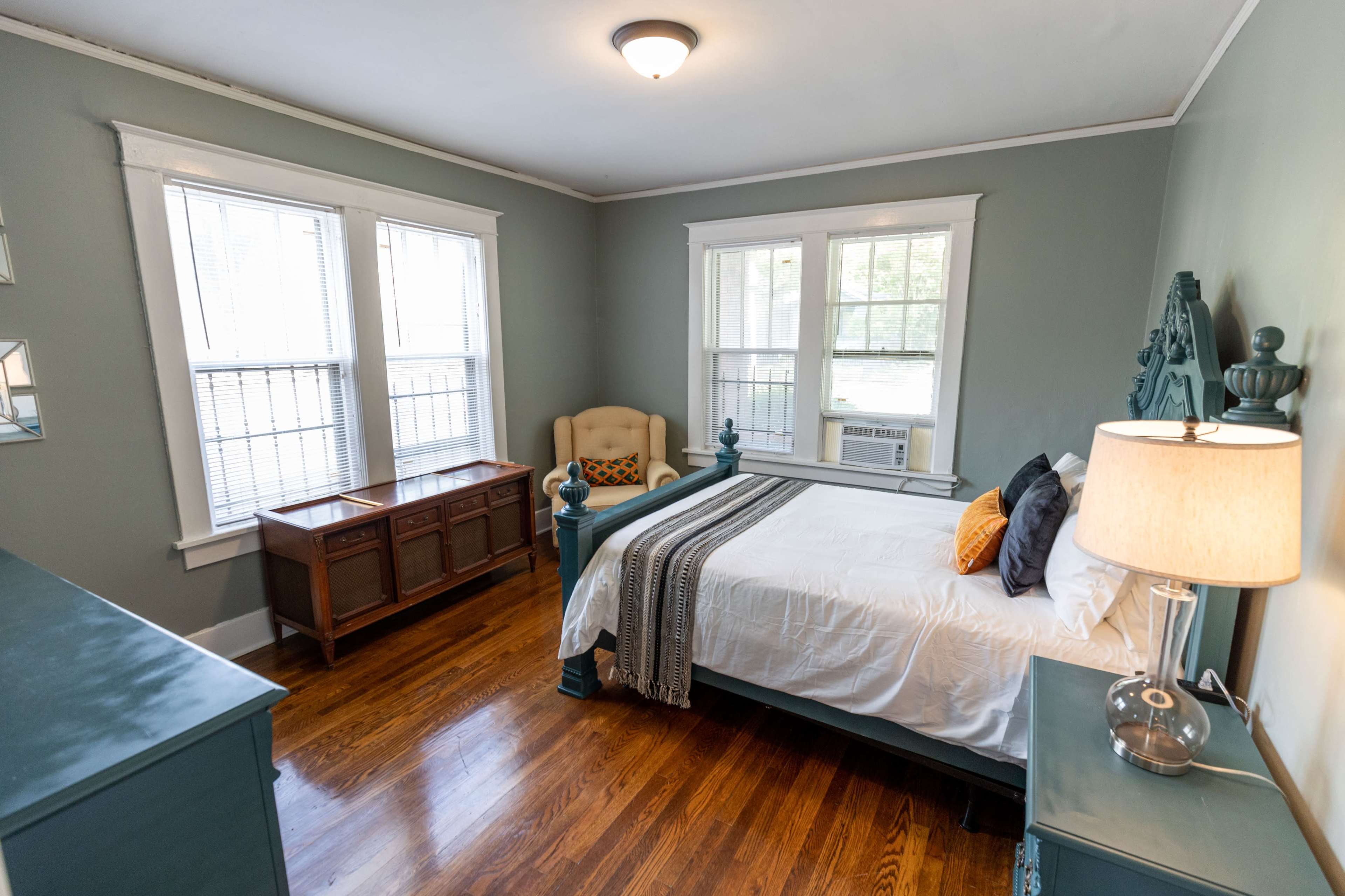 A bedroom with a bed, an armchair, a dresser, and two large windows letting in natural light.