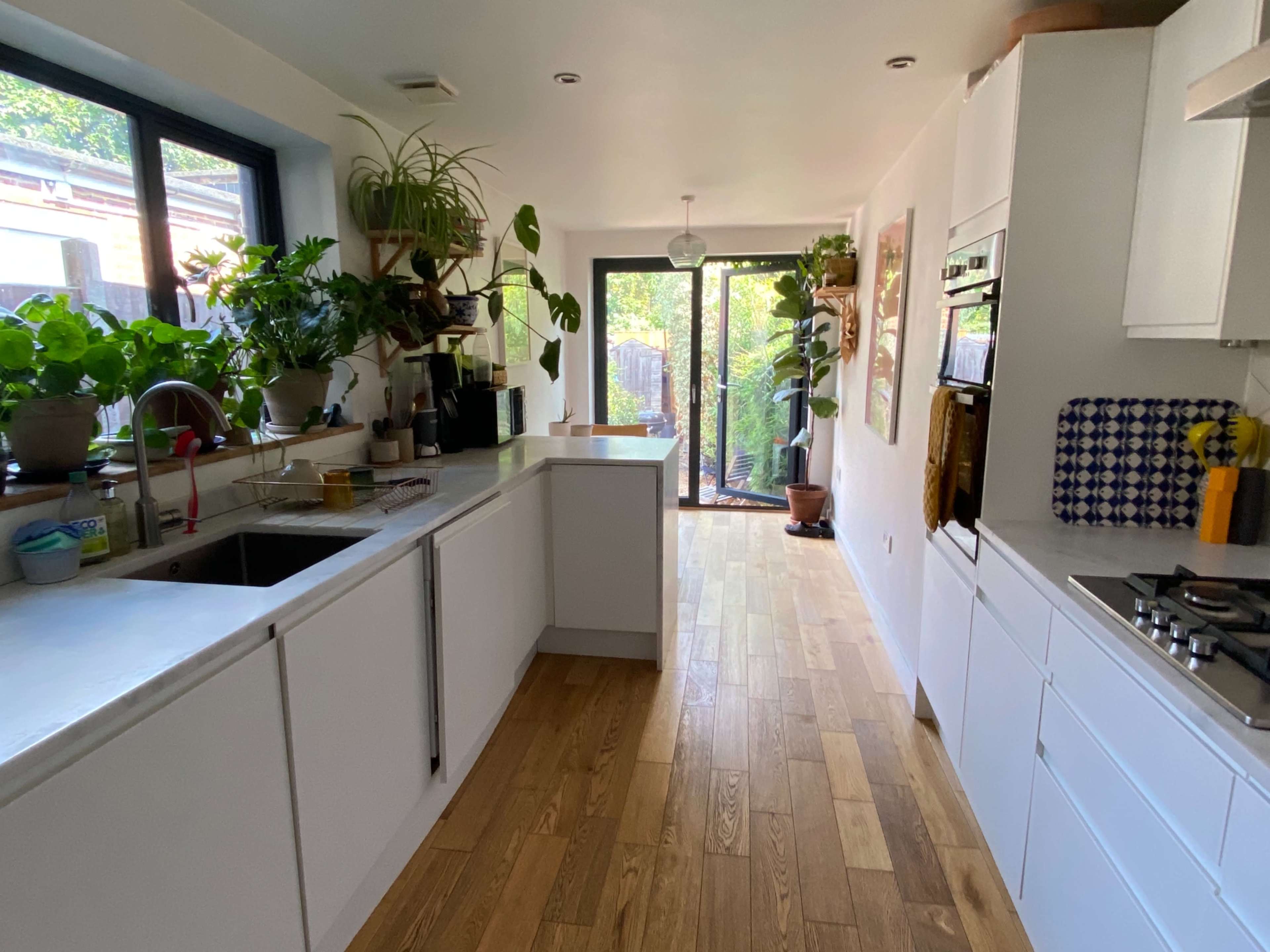 A modern kitchen with white cabinetry, wooden flooring, and large windows facing a garden filled with plants.